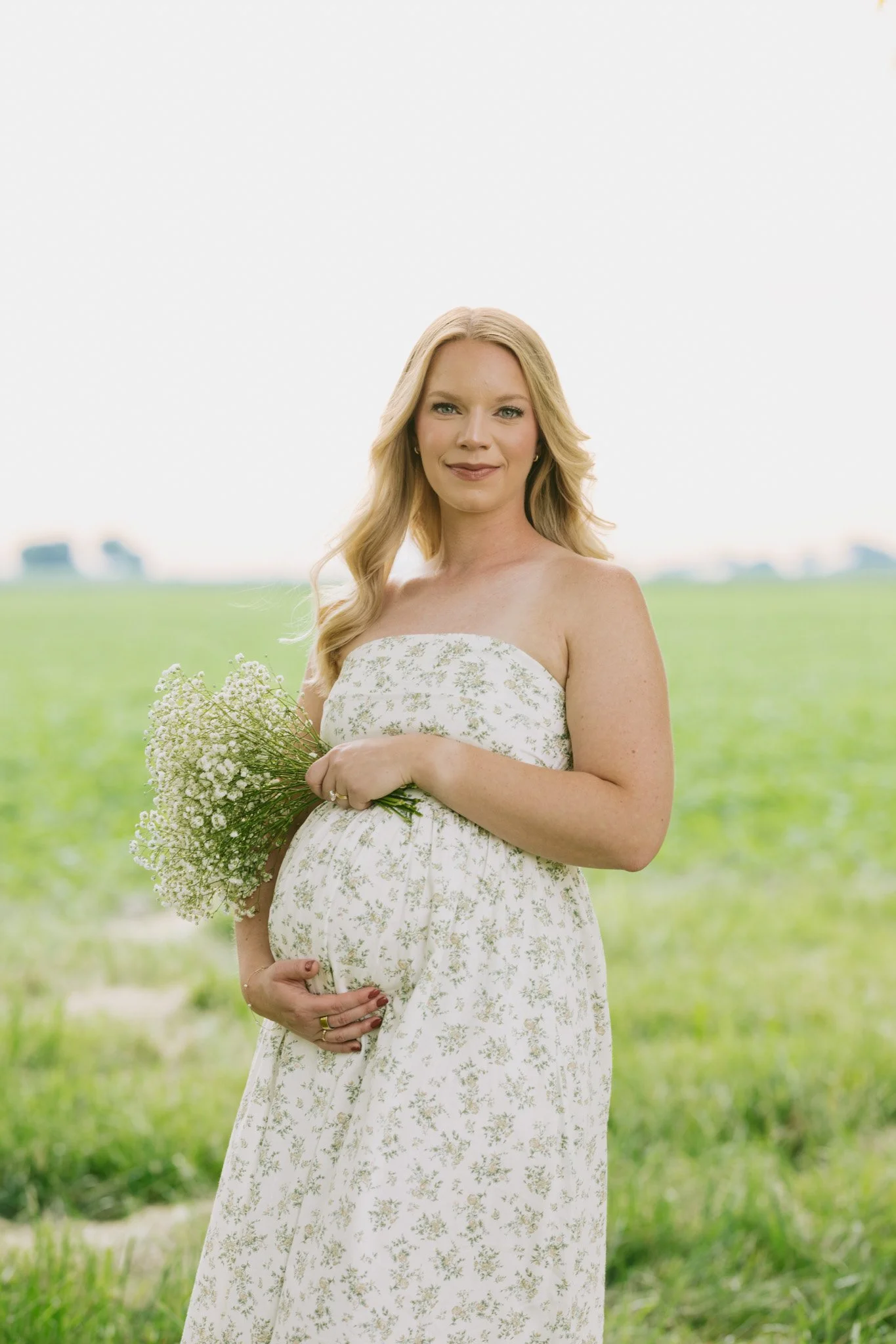 Pregnant woman in a floral dress standing outdoors in a green field, holding a bouquet of small white flowers, with long blonde hair and a gentle smile.