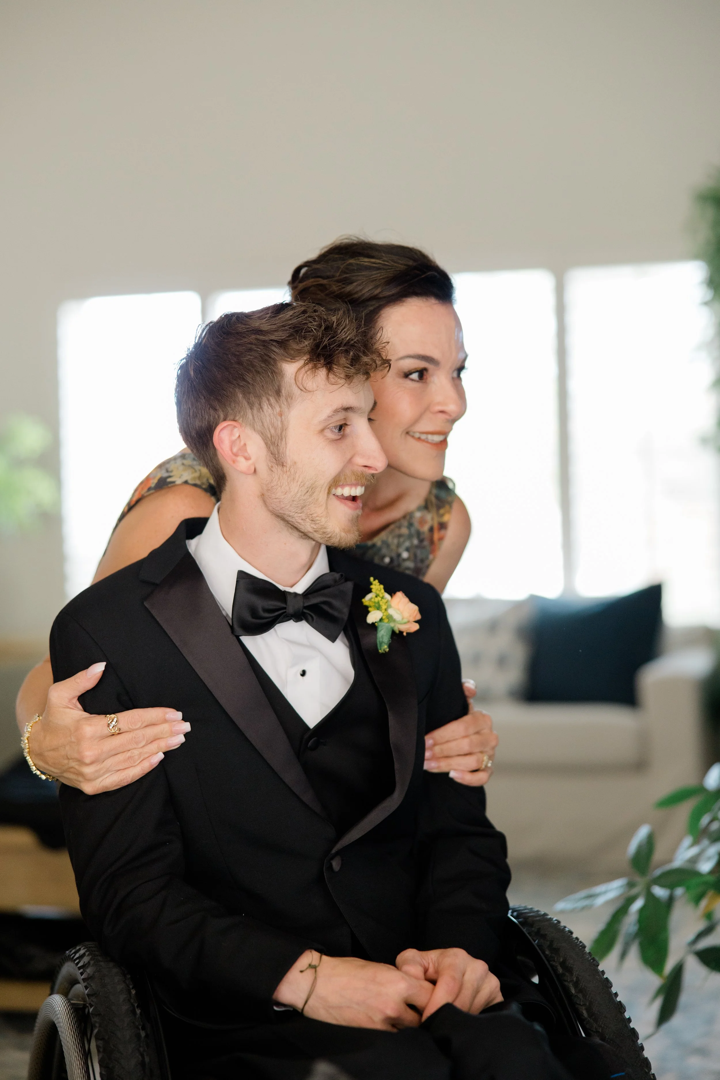 A young man in a wheelchair dressed in a black tuxedo with a bow tie, smiling, being embraced from behind by a woman with dark hair in a floral dress, smiling, in a well-lit room with large windows and garden plants.
