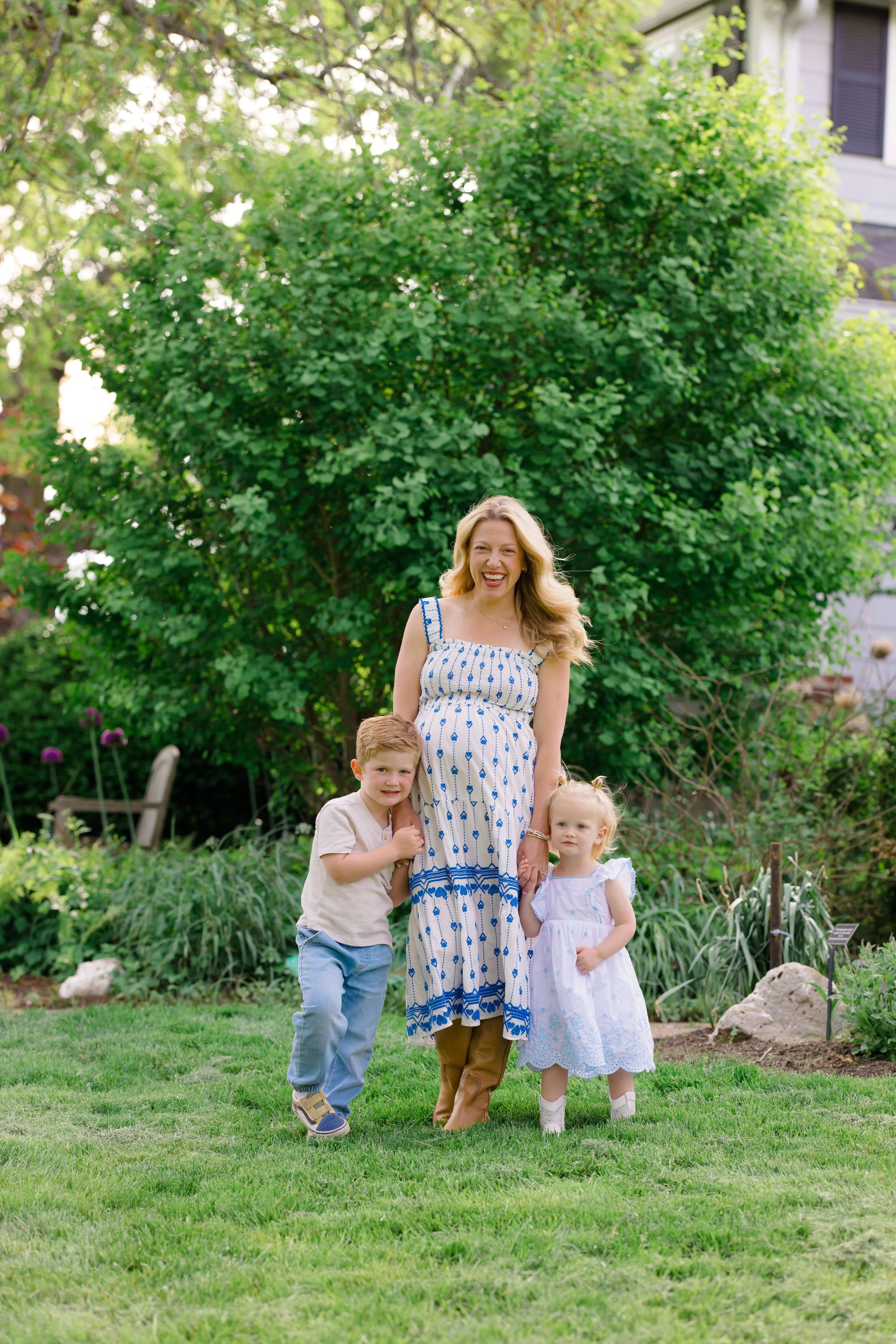 A woman with long blonde hair in a dress with blue and white pattern standing outside in front of a large green bush, holding hands with a young girl in a white dress, and surrounded by a green garden with two young boys, all smiling and posing for t
