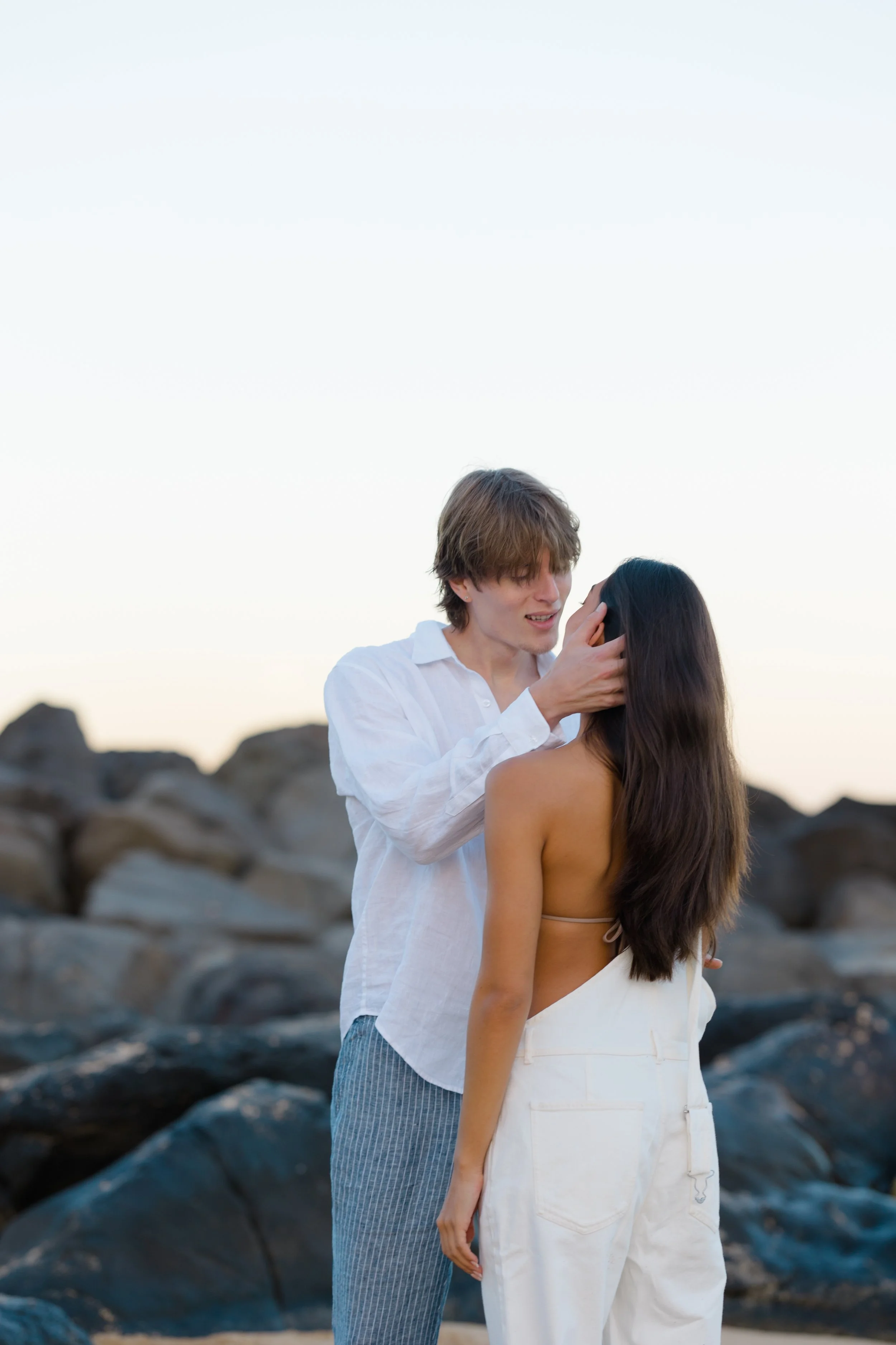 A young man holding a woman's face and looking into her eyes on a rocky beach at sunset.