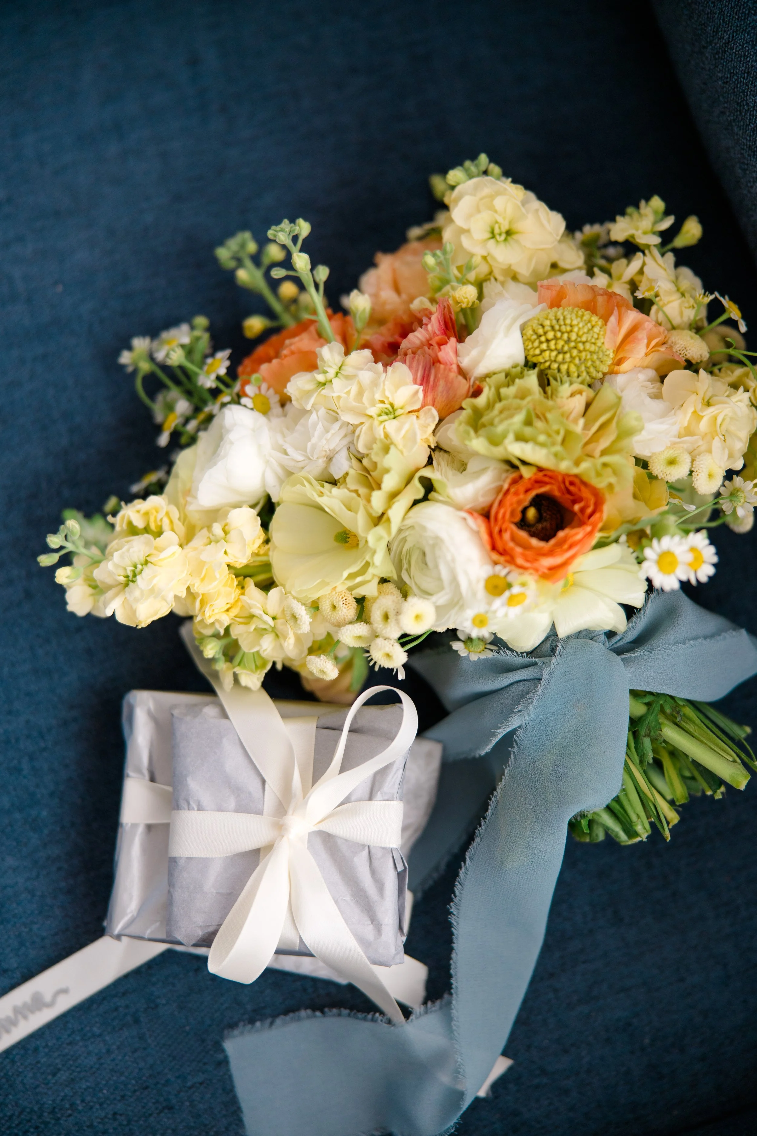 A bouquet of mixed flowers with white, peach, and yellow blooms, wrapped in blue paper with a white ribbon, resting on a dark textured surface.