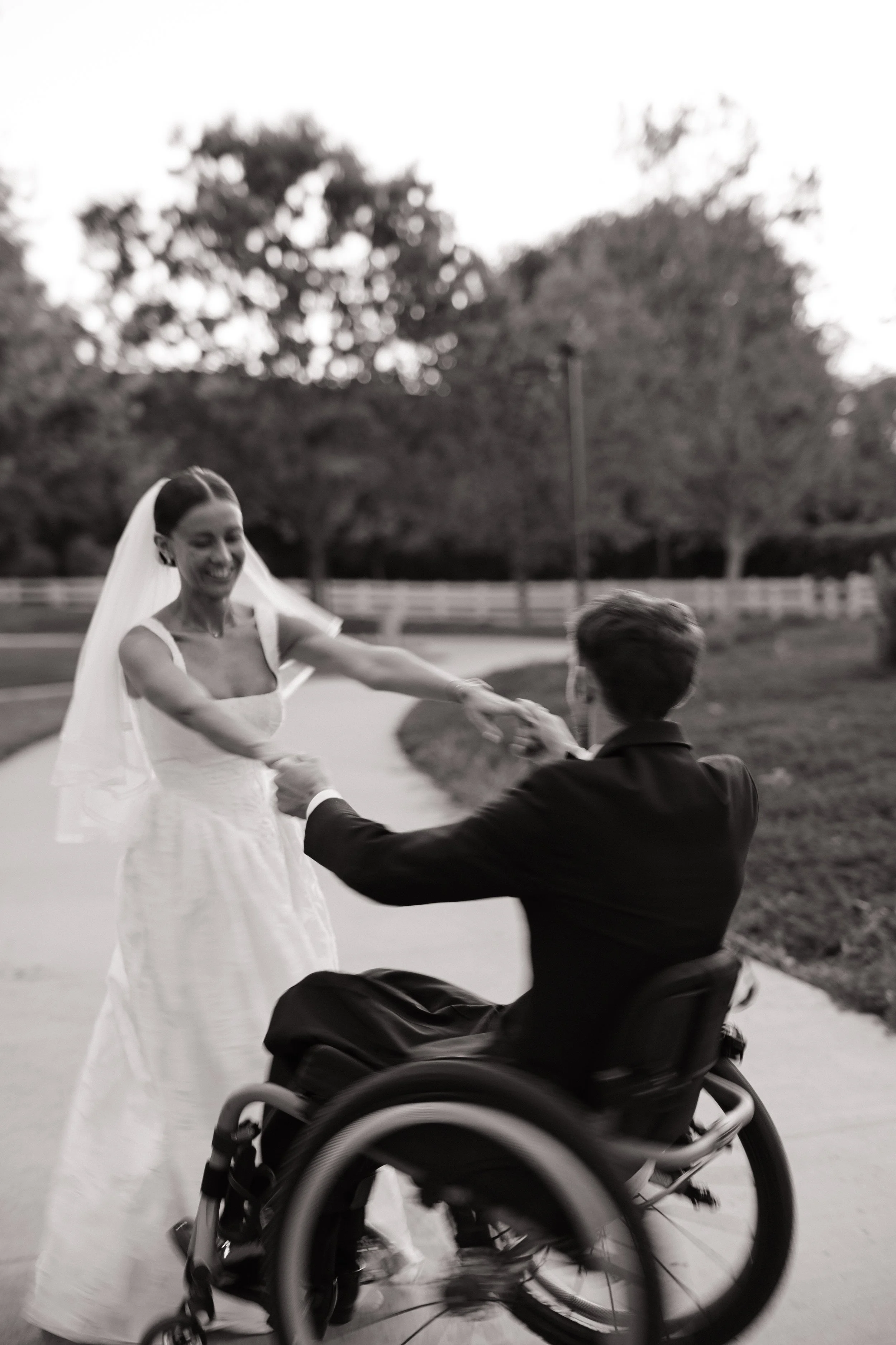 A woman in a wedding dress, wearing a veil, is smiling and holding hands with a man in a wheelchair. They are outdoors with trees and a fence in the background.
