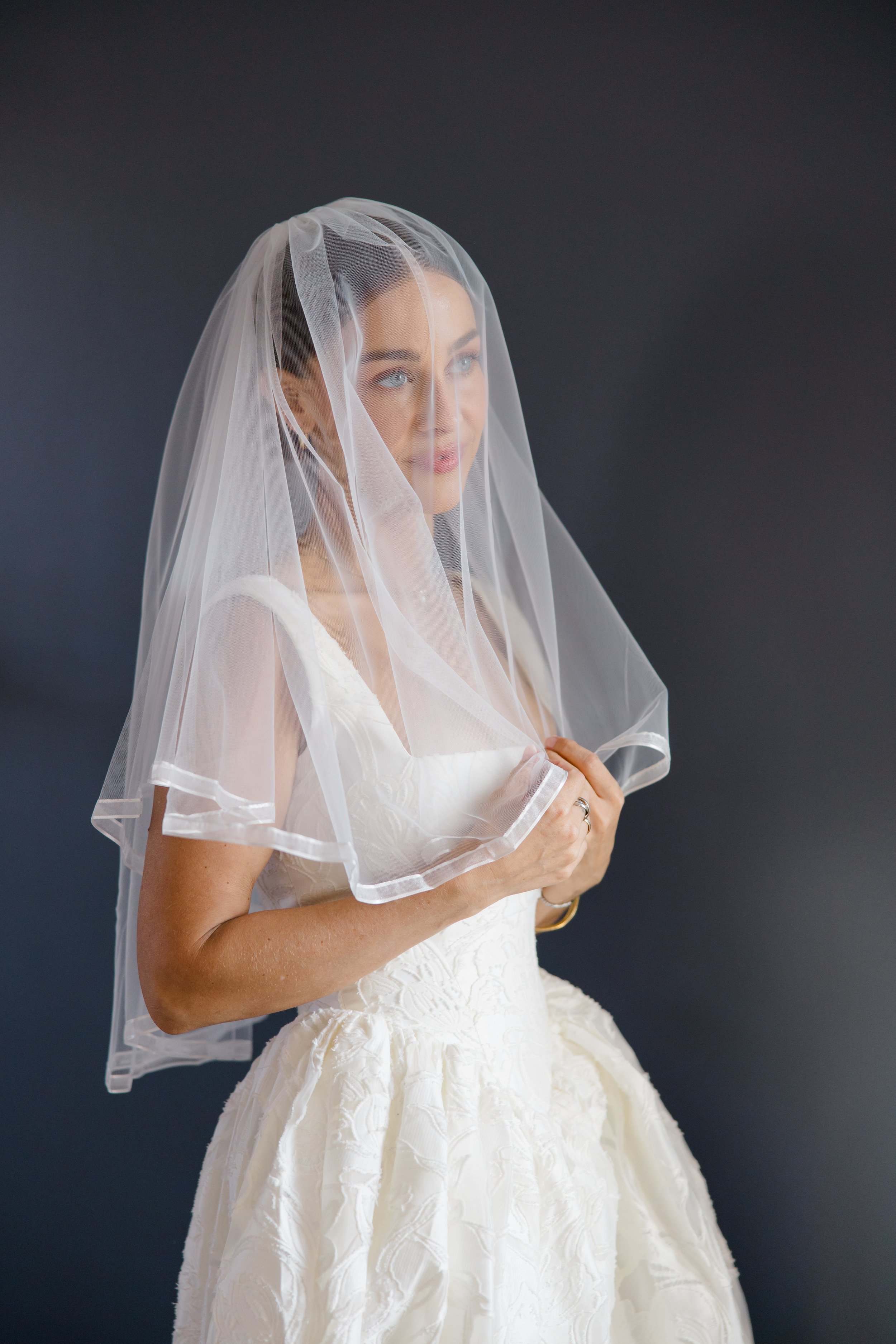 A woman in a white wedding dress with a sheer veil over her face, standing against a dark background.