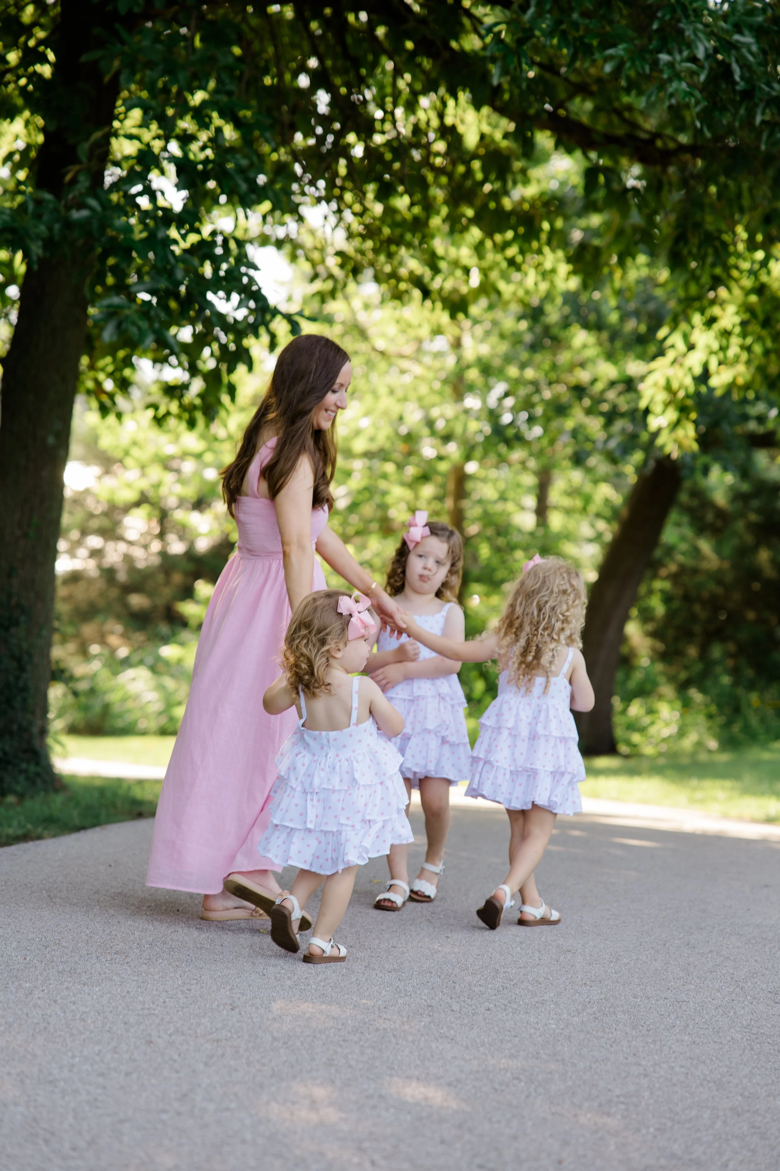 A woman in a pink dress walking with four young girls dressed in white polka dot dresses with pink bows, outdoors in a park with tall trees and sunshine.