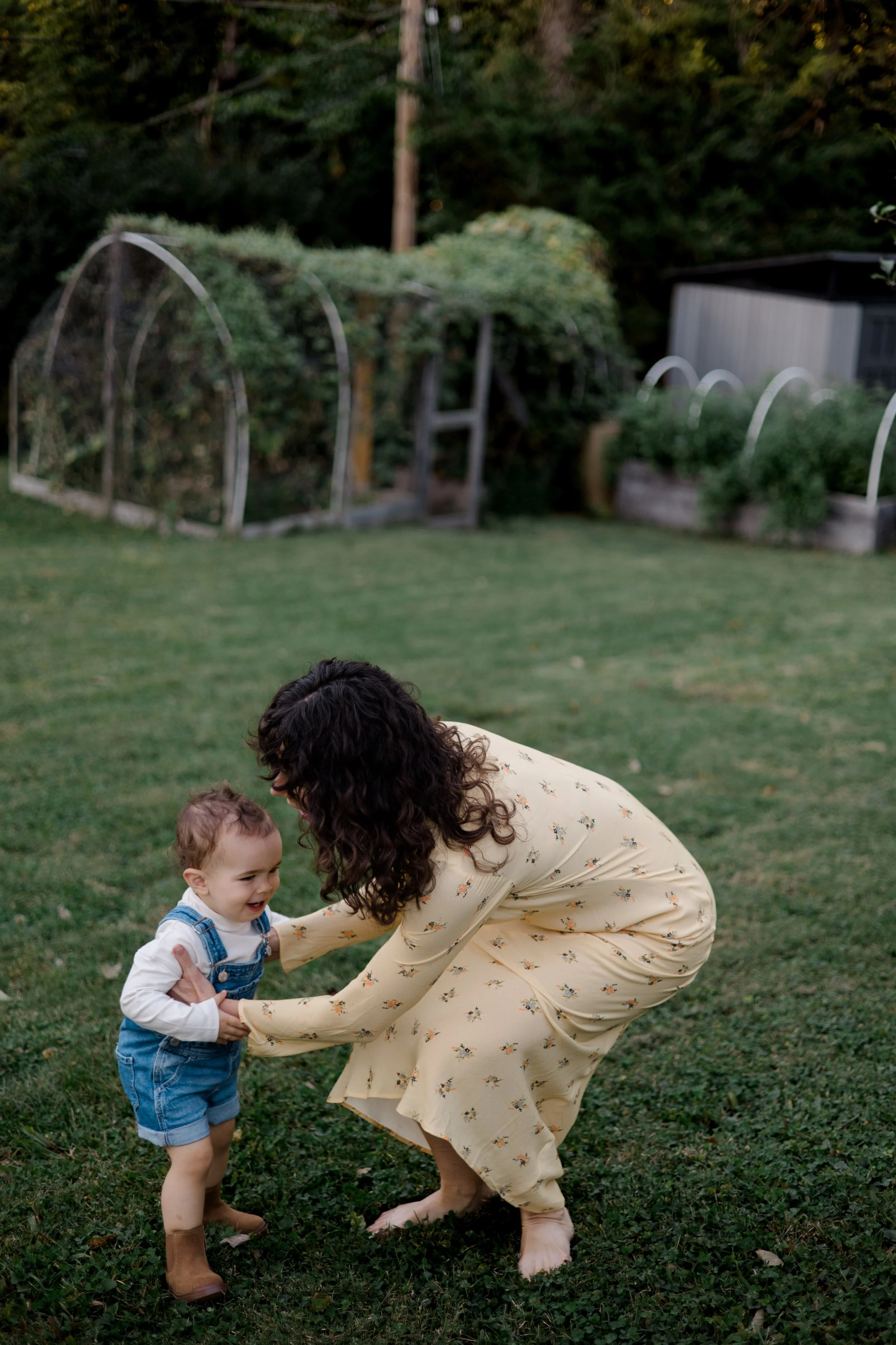 A woman with curly hair in a yellow dress playing with a young boy in a grassy yard, holding his hands as they smile and laugh.