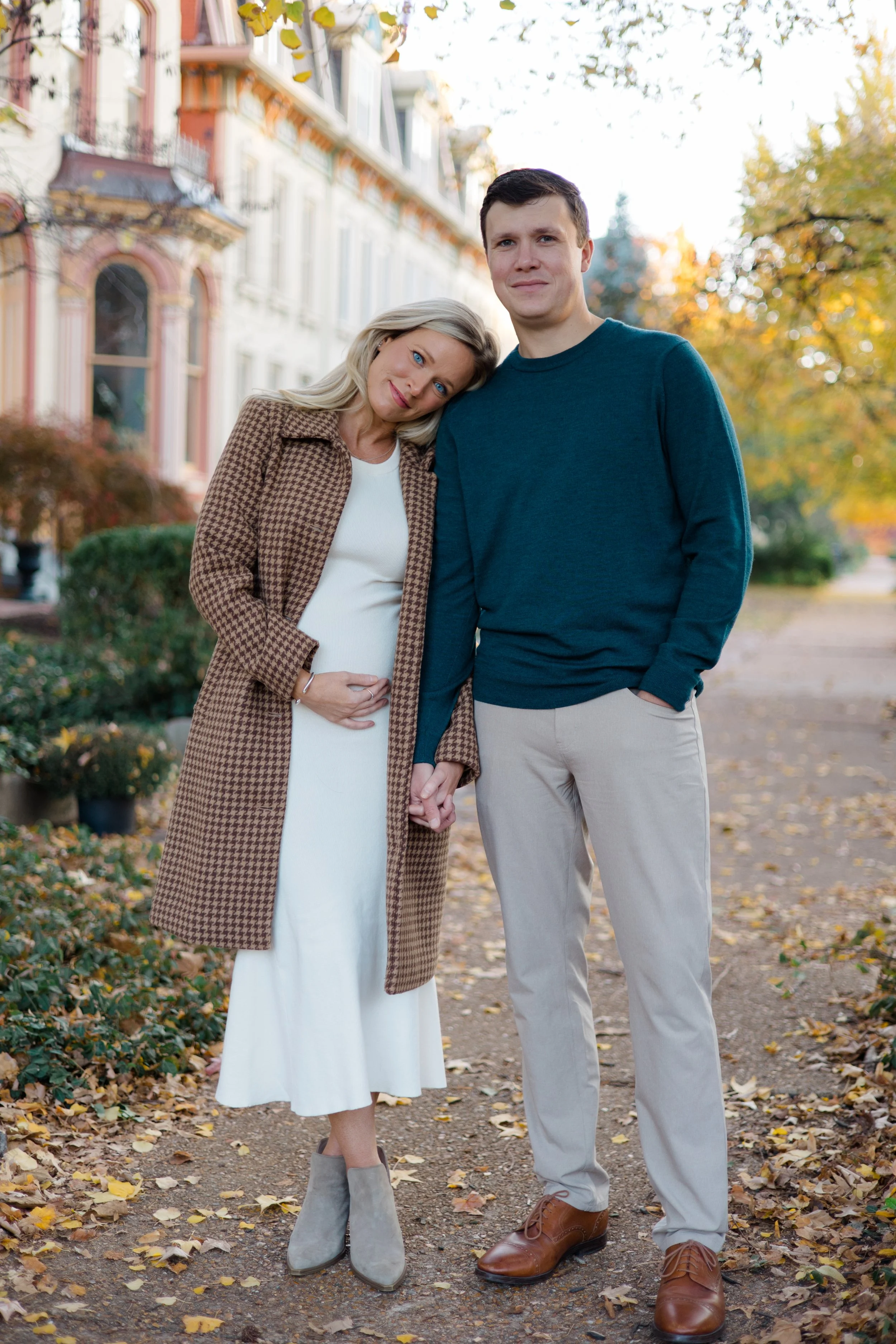 A pregnant woman and a man stand together outdoors on a fall day, holding hands. The woman rests her head on the man's shoulder, and they are surrounded by trees with yellow and orange leaves, with fallen leaves on the ground. The woman wears a brown