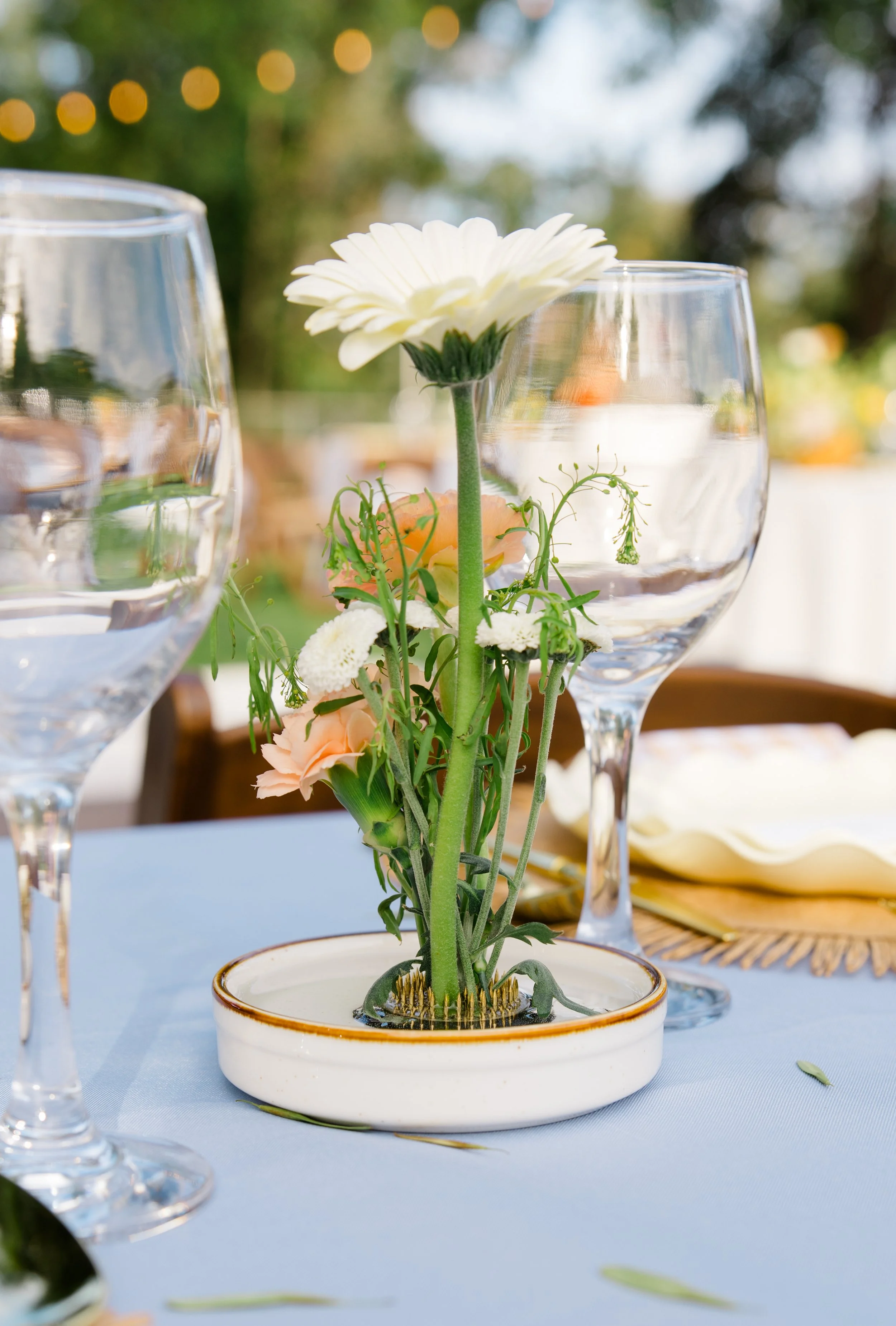 A white flower in a small ceramic pot on a table, with two empty wine glasses and blurred outdoor background.