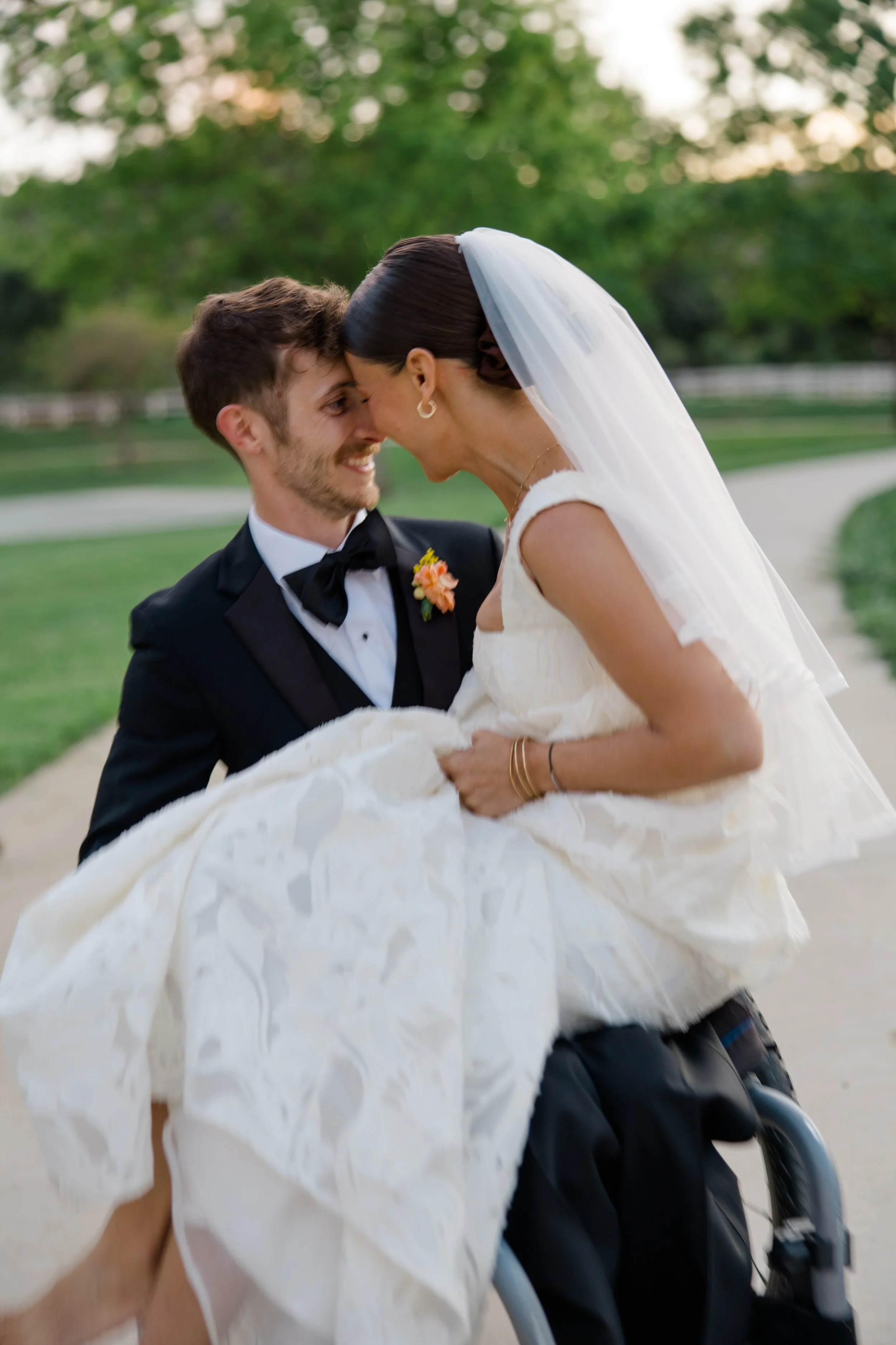 A bride in a white wedding dress and veil is being held by a man in a wheelchair, dressed in a tuxedo with a boutonniere, outdoors with trees in the background.