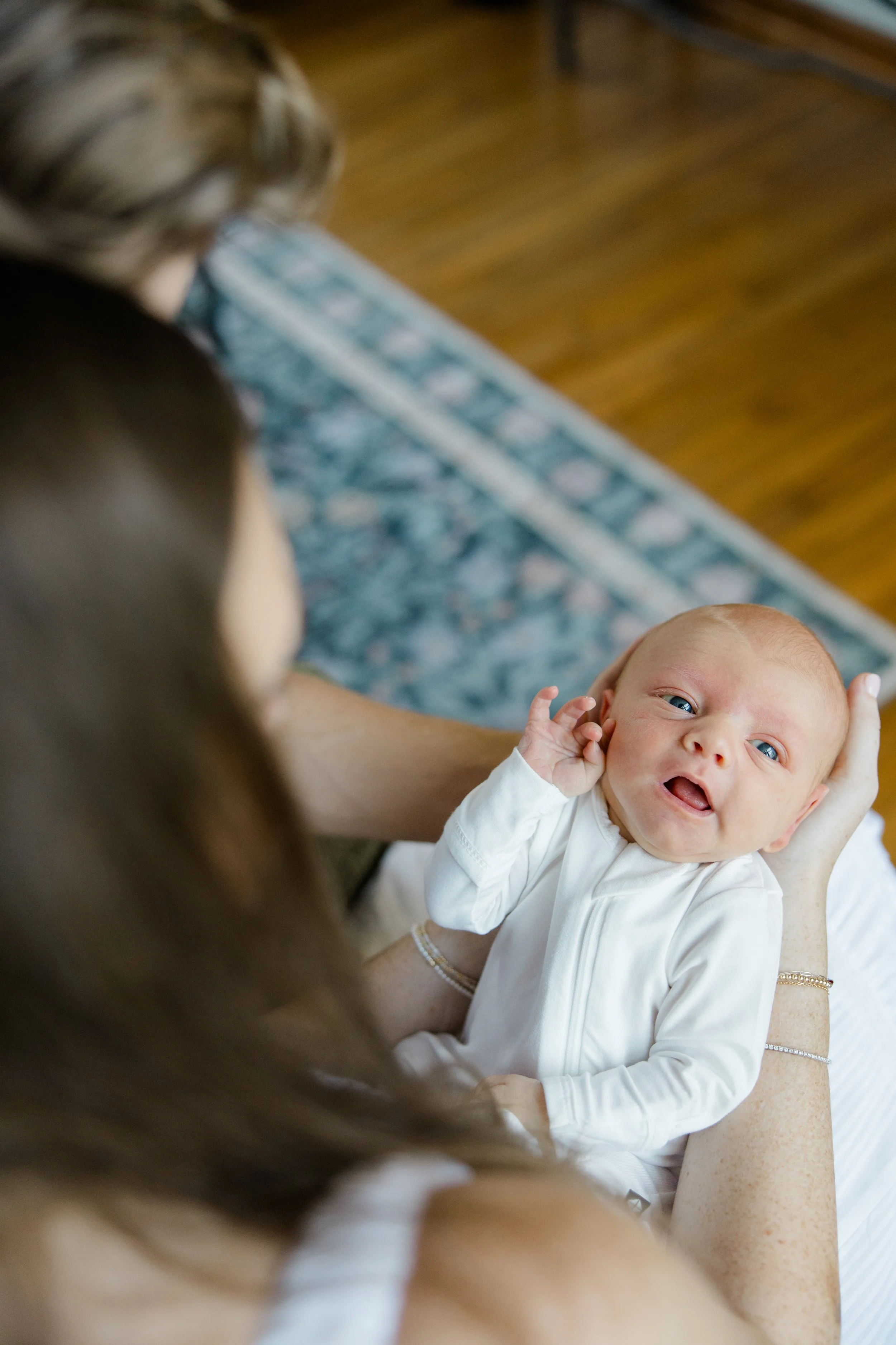 A baby with blue eyes and red hair looking up and smiling while being held by a woman.
