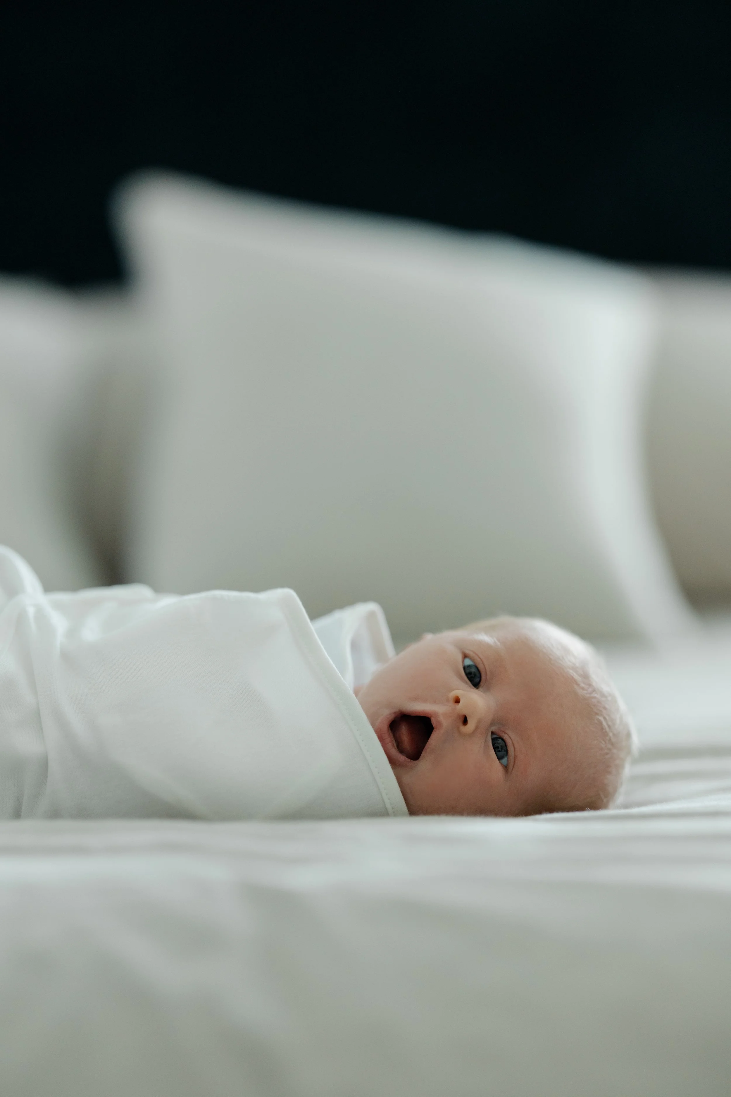 A newborn baby laying on a white blanket, wearing a white outfit, with eyes open and mouth slightly open, in a hospital or medical setting.