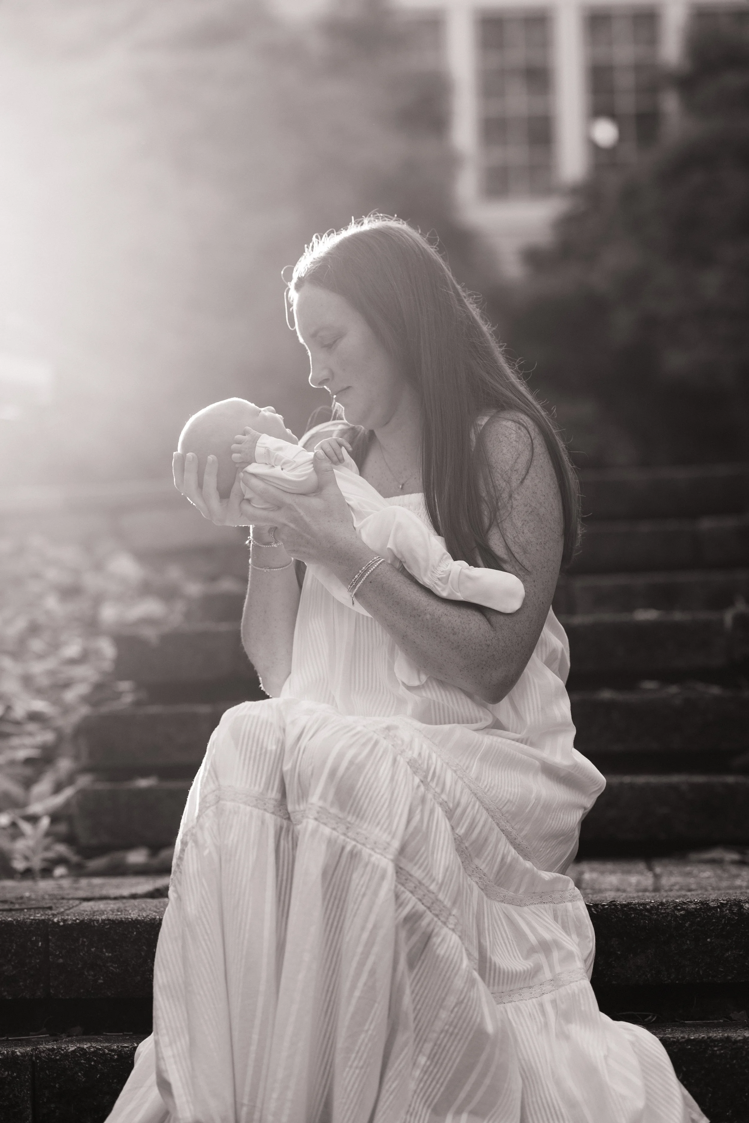 A woman with long hair wearing a white dress is sitting on stairs outdoors, holding a small baby in her hands and gently kissing the baby.
