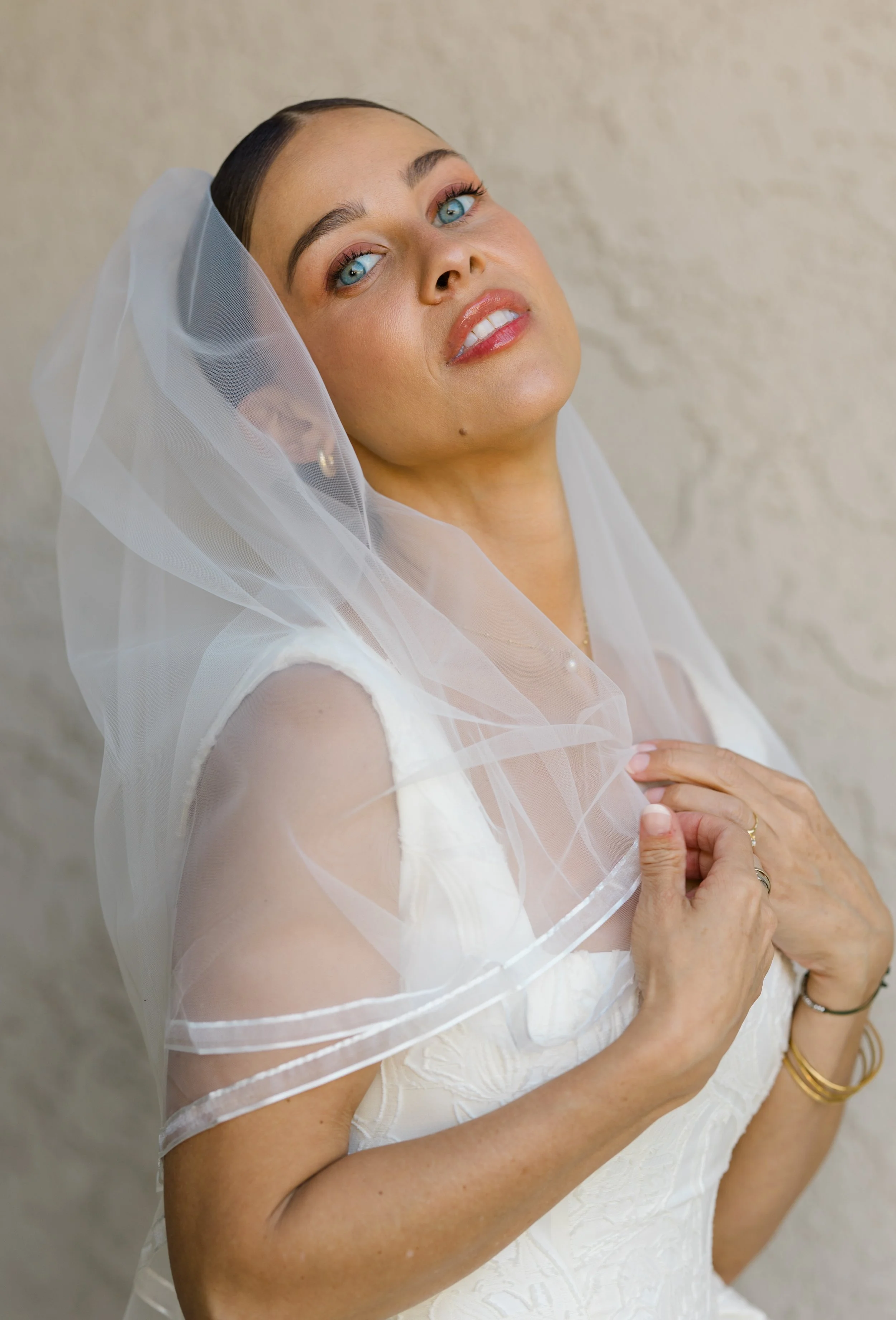 A woman with blue eyes and dark hair tied back, wearing a white dress and veil, looking at the camera with a gentle expression.