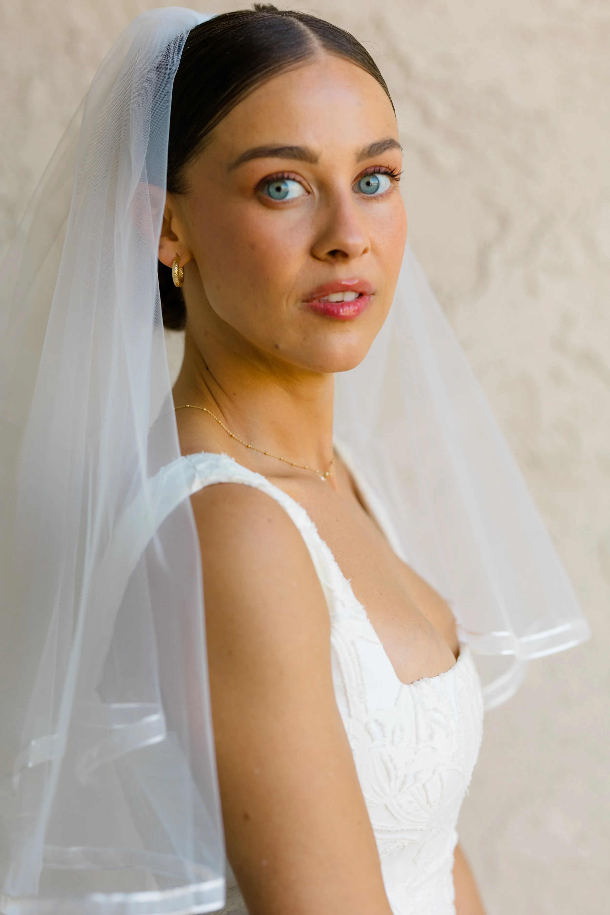 Close-up of a young woman with blue eyes wearing a white dress, gold hoop earrings, and a delicate gold necklace, with a white veil on her head, near a beige wall.