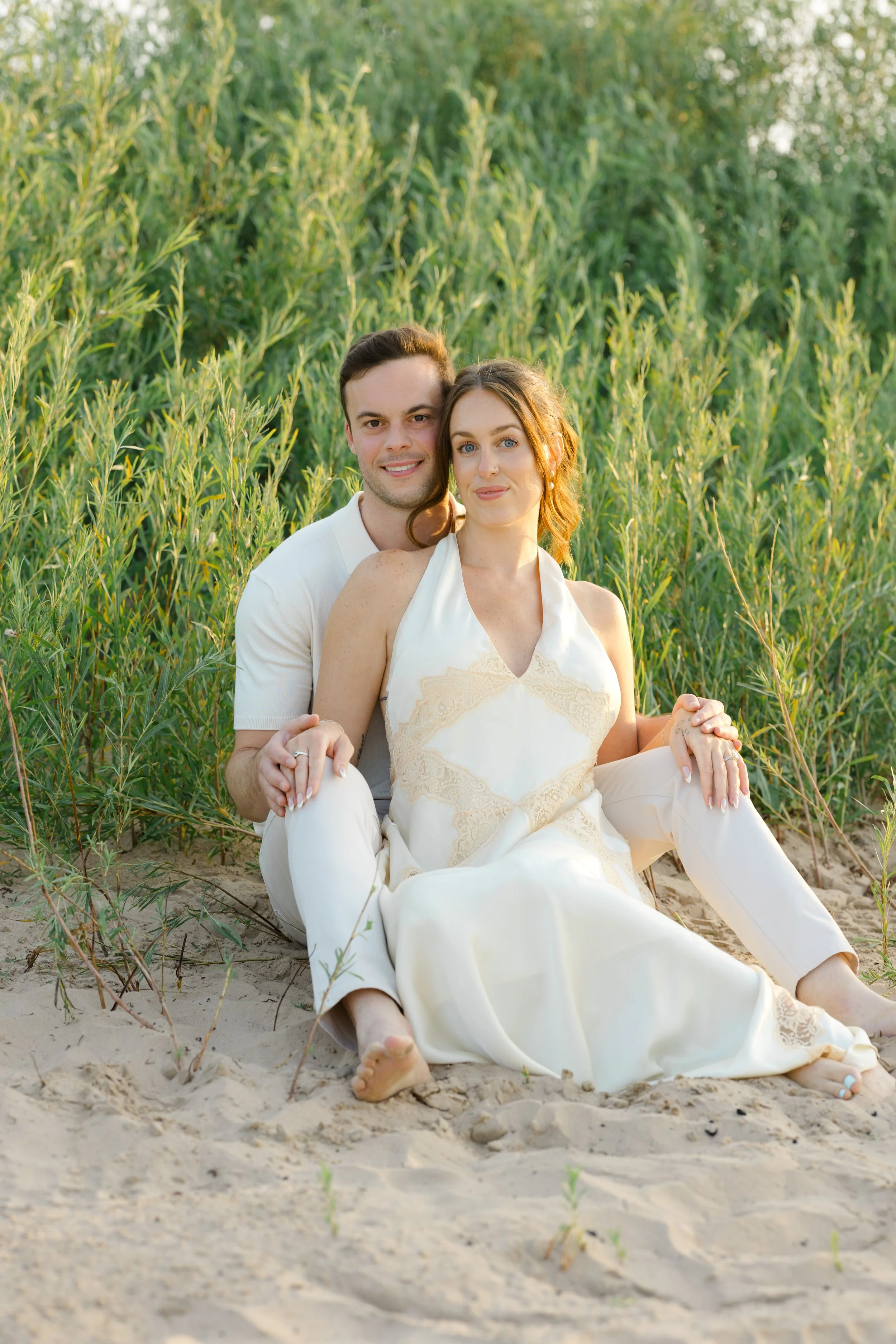 A couple sitting on the sandy ground in front of green plants, smiling with the man behind the woman, both dressed in white, during sunset.