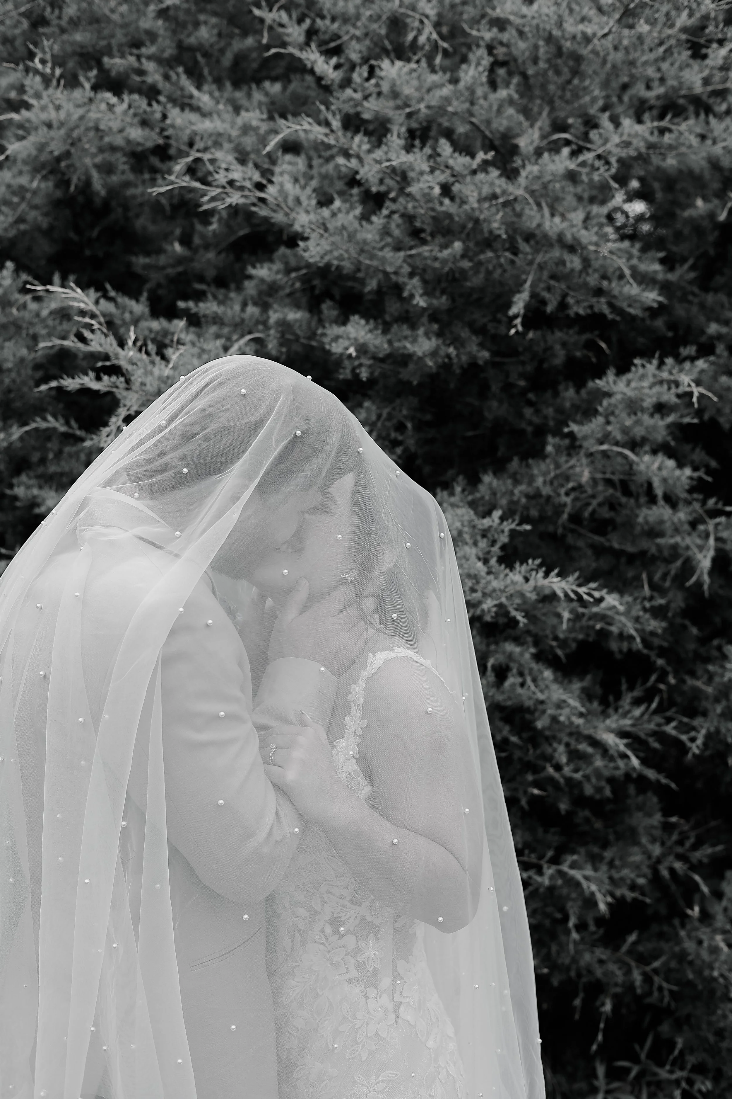 A person wearing a wedding dress and veil, standing outdoors in front of dense, leafy trees.