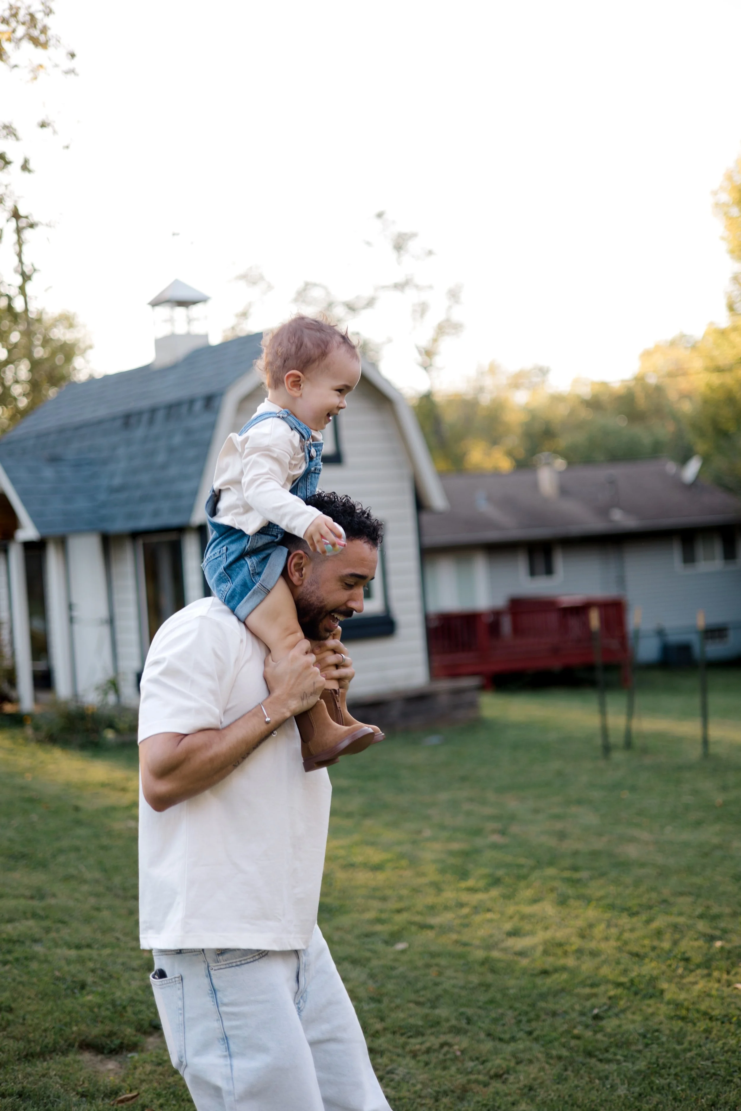 A man with a beard and light blue jeans is carrying a smiling young child on his shoulders in a backyard with green grass, trees, and houses in the background during sunset.