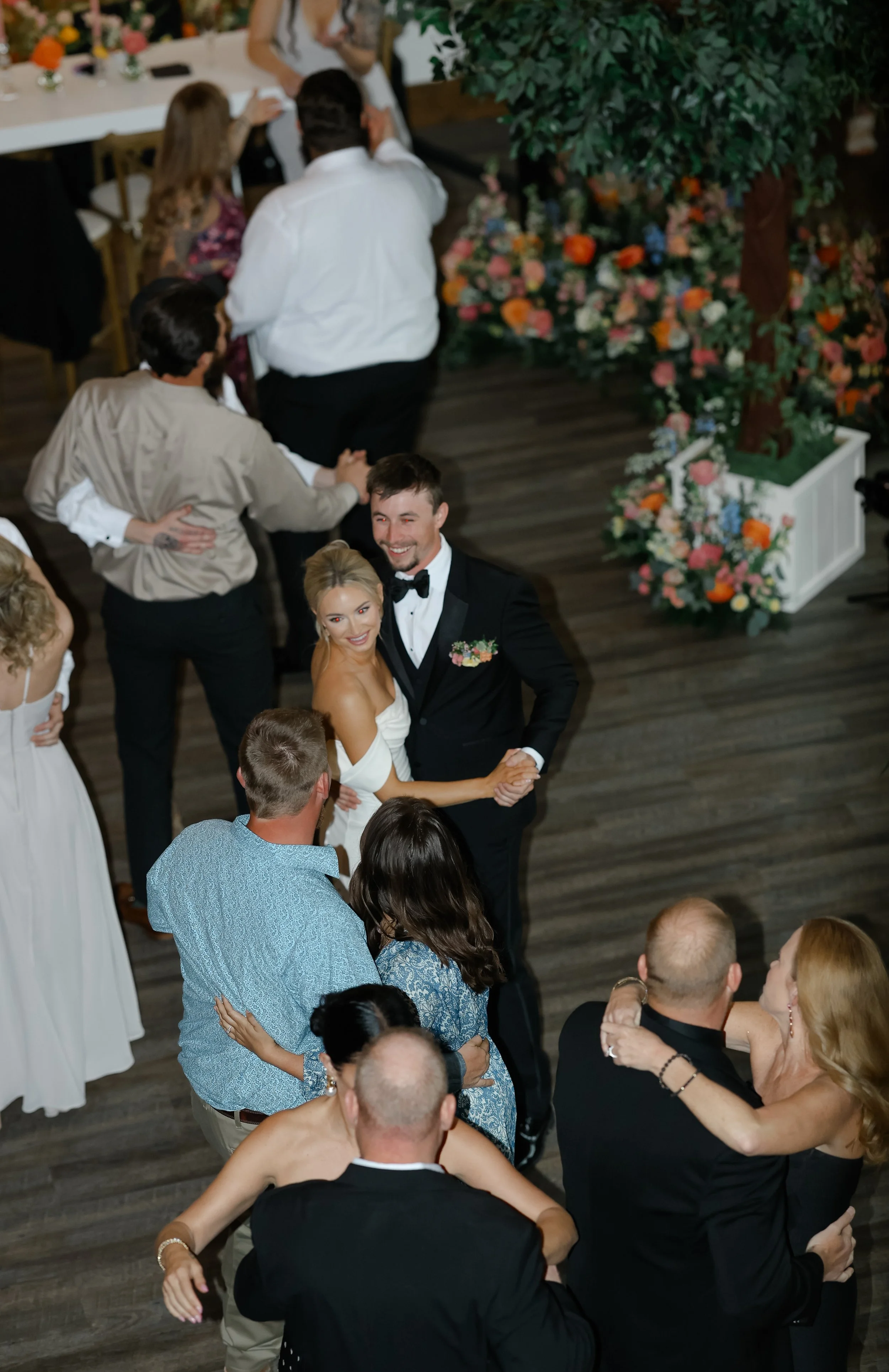 A bride and groom dancing together at their wedding reception, surrounded by guests, in a decorated venue with floral arrangements and trees.