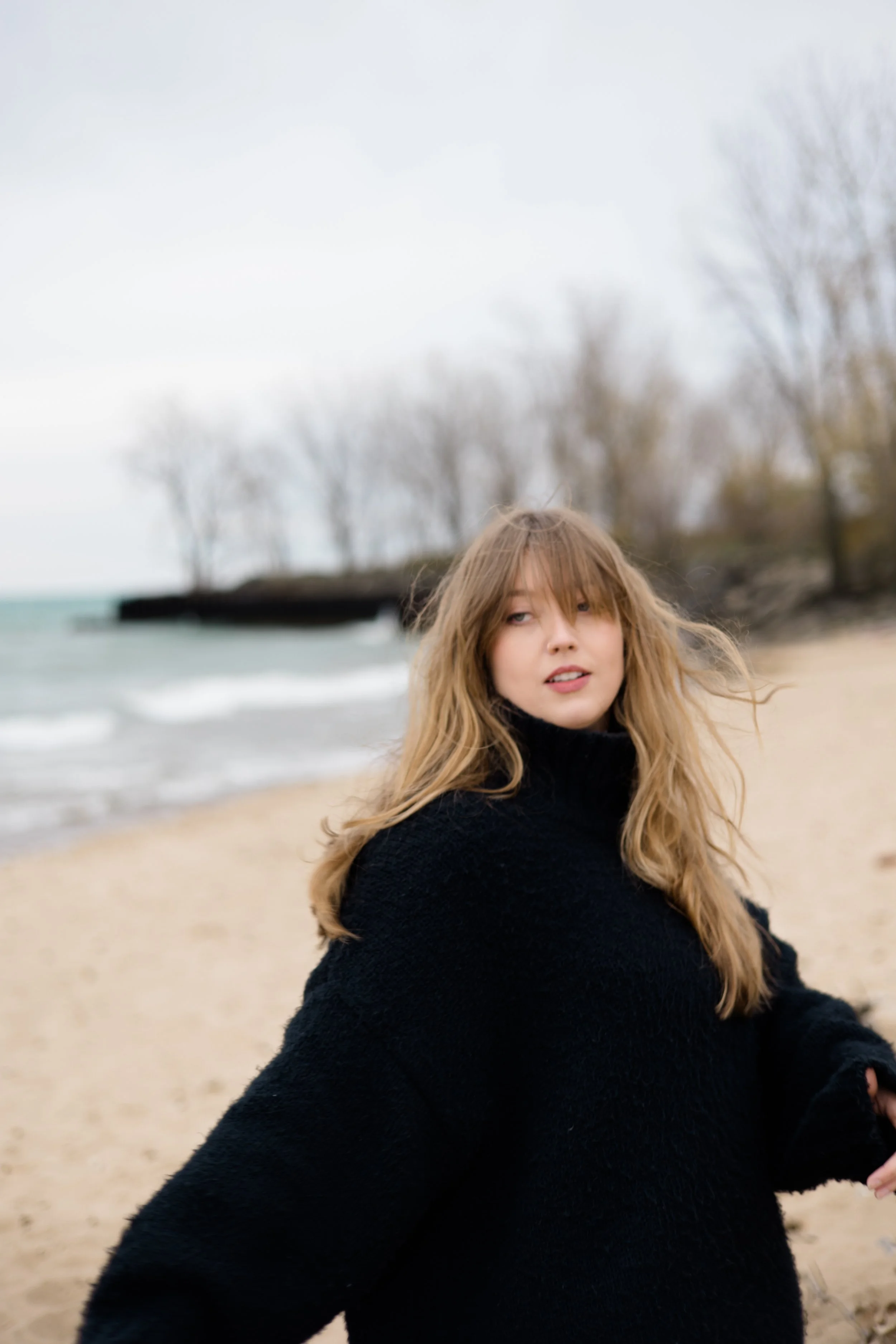 A young woman with long blonde hair and bangs, wearing a black sweater, standing on a sandy beach with leafless trees and a cloudy sky in the background.
