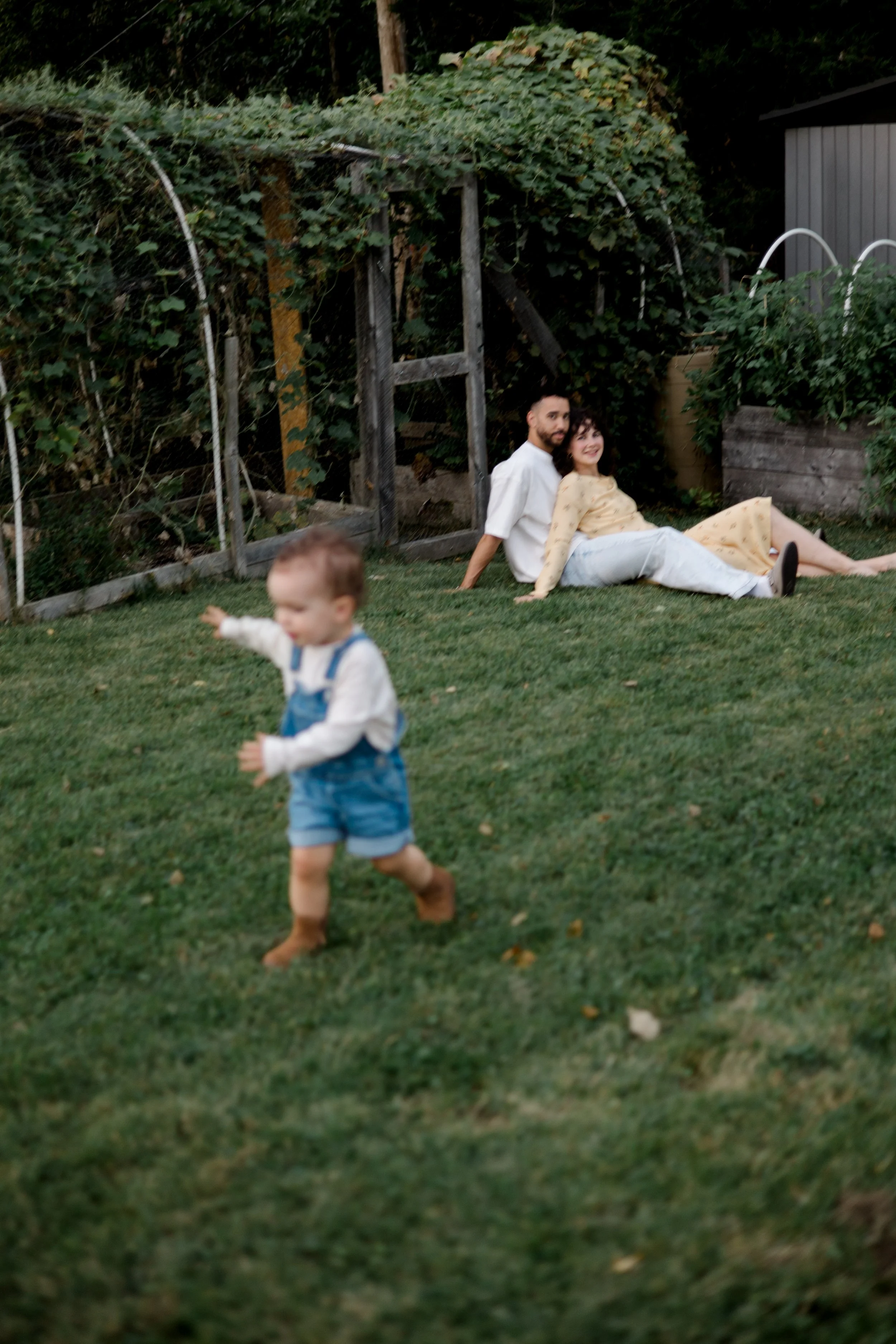 A couple sitting on grass in a backyard, with a child running in the foreground.