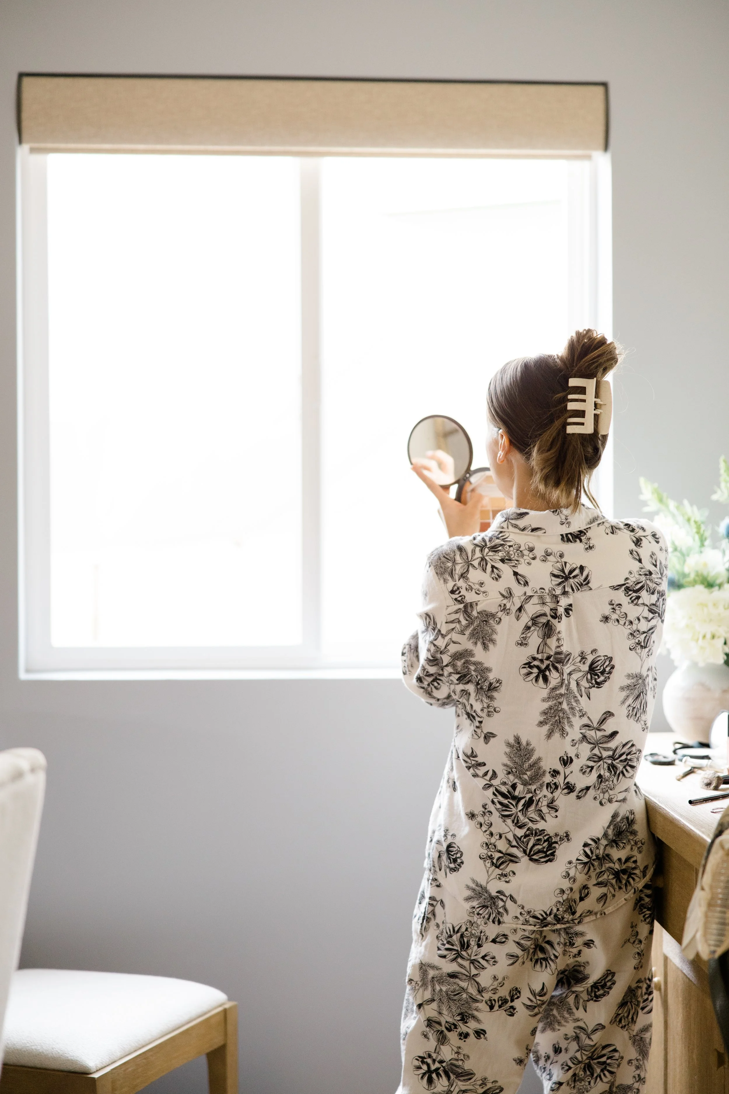 A woman with hair in a bun held with a large beige hairclip is standing indoors, facing a window, holding a round mirror in her right hand, wearing a floral print outfit with branches and leaves.