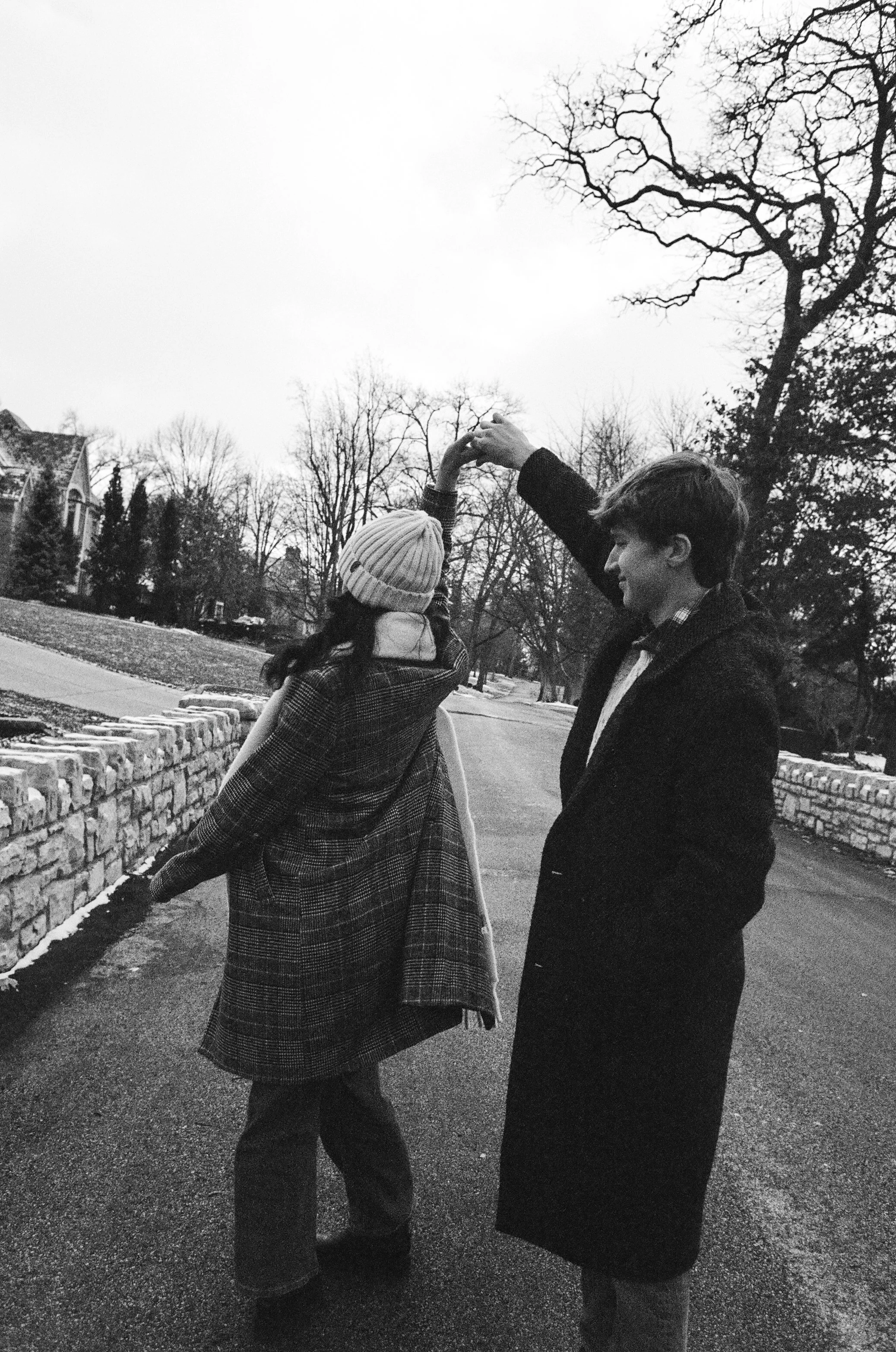 Two young people dancing together outdoors in a park on a cold day. The girl is wearing a knit hat and a long plaid coat, while the boy is wearing a dark jacket. They are holding hands and smiling amid bare trees and a stone wall.