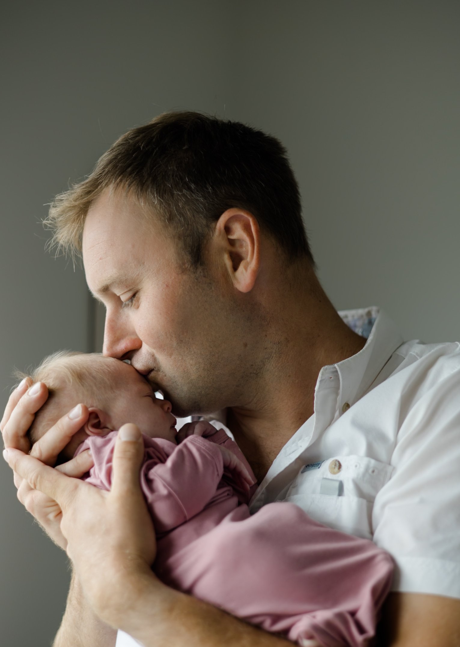 A man with short brown hair and wearing a white shirt is holding a newborn baby with blond hair and pink clothing, closely kissing the baby's forehead while the baby sleeps peacefully.