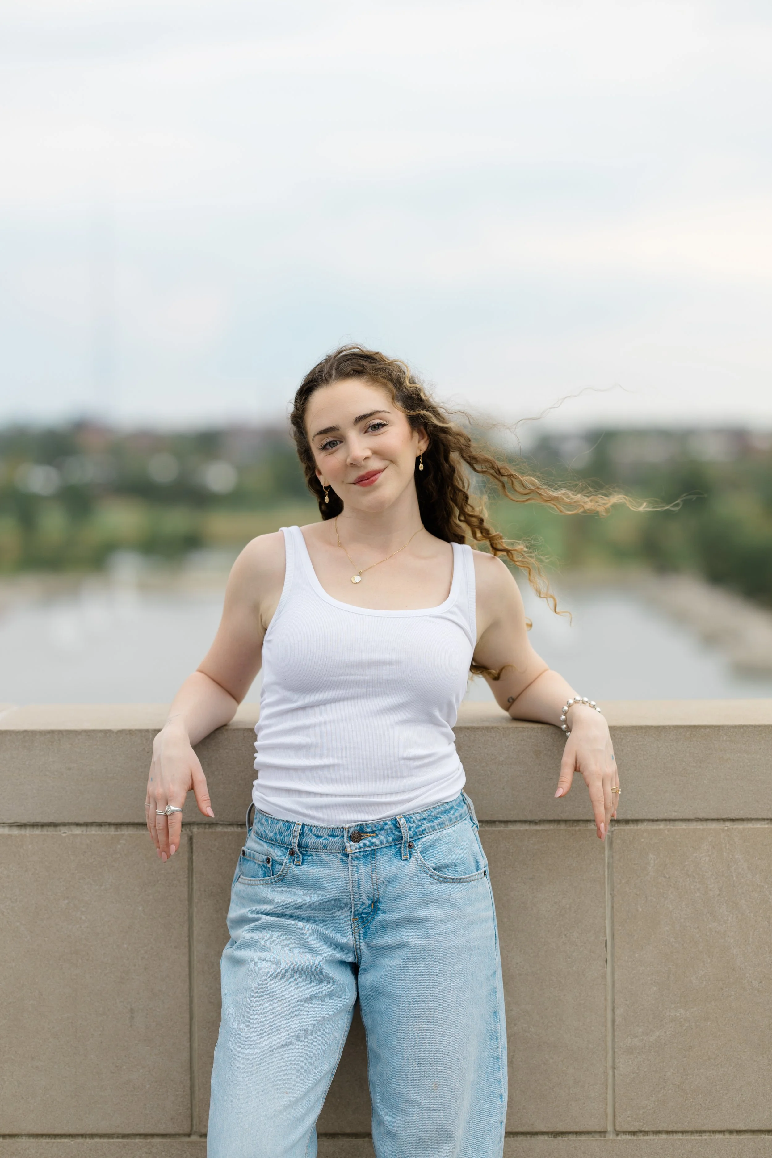 A young woman with curly hair wearing a white tank top and light blue jeans standing outdoors near a concrete railing, with a blurred cityscape and river in the background.