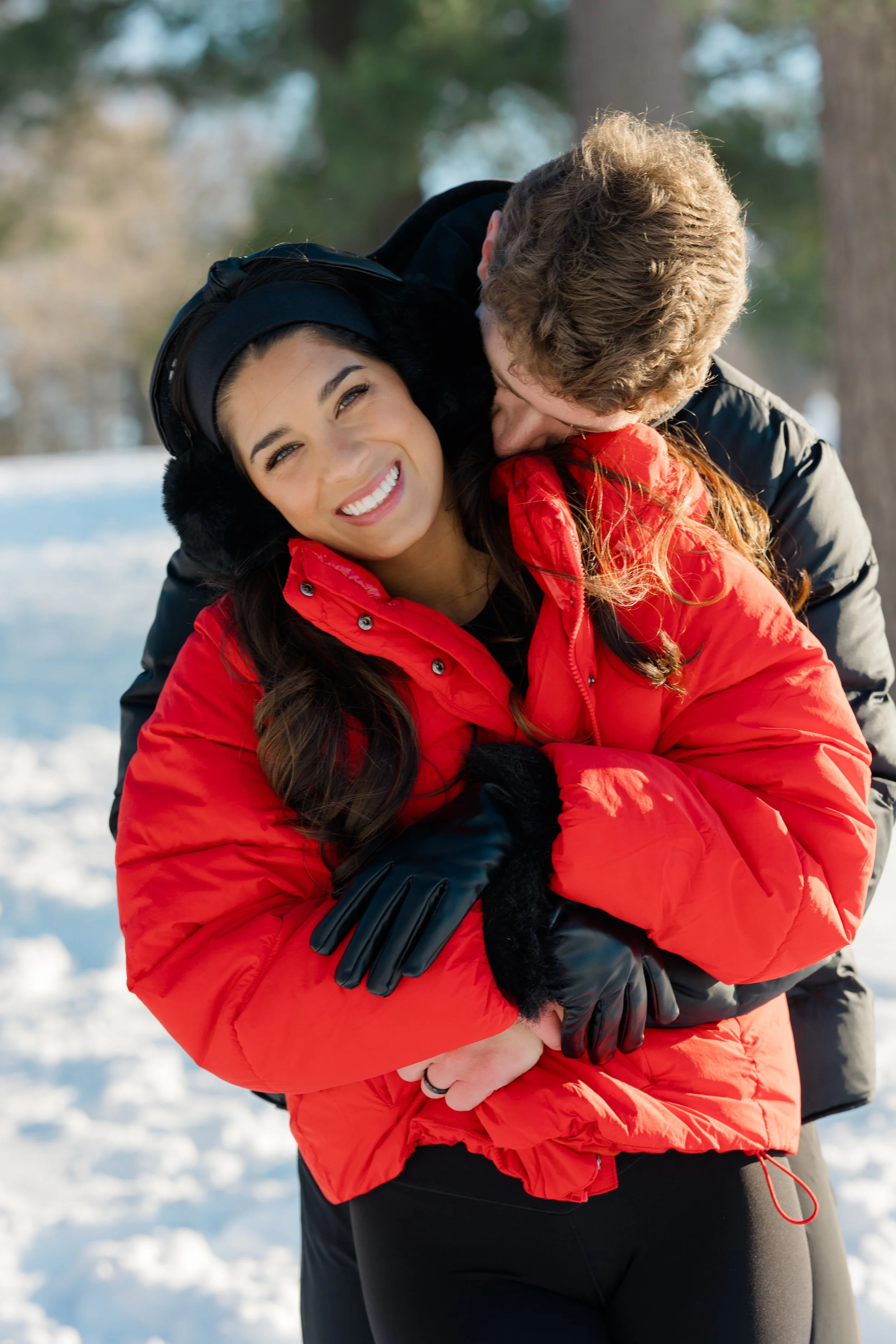 A smiling woman in a red winter jacket and black gloves being hugged by a person in a black jacket outdoors in a snowy park with trees.