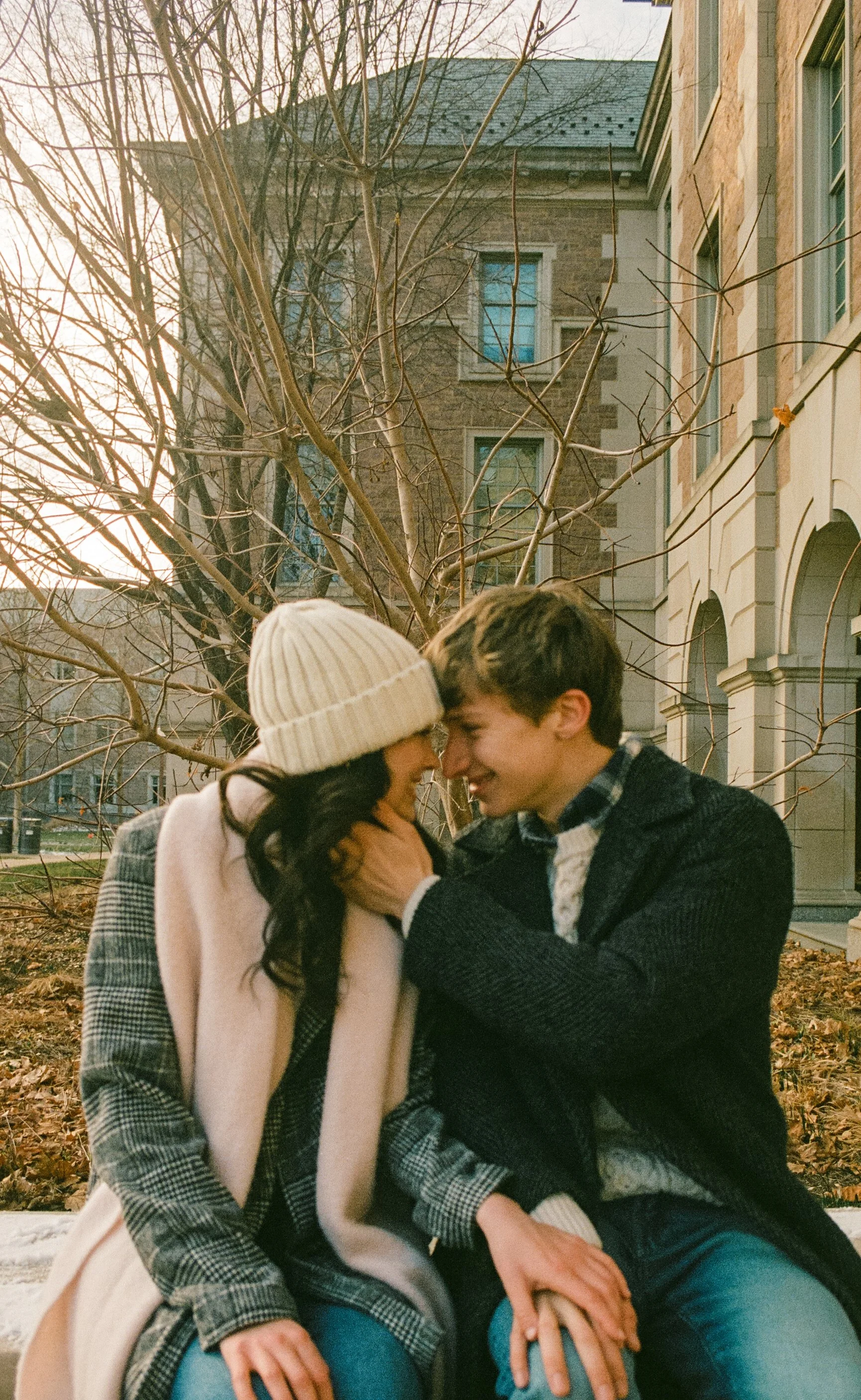 A couple sitting close together outdoors, sharing a joyful moment, with a leafless tree and buildings in the background during fall or winter.