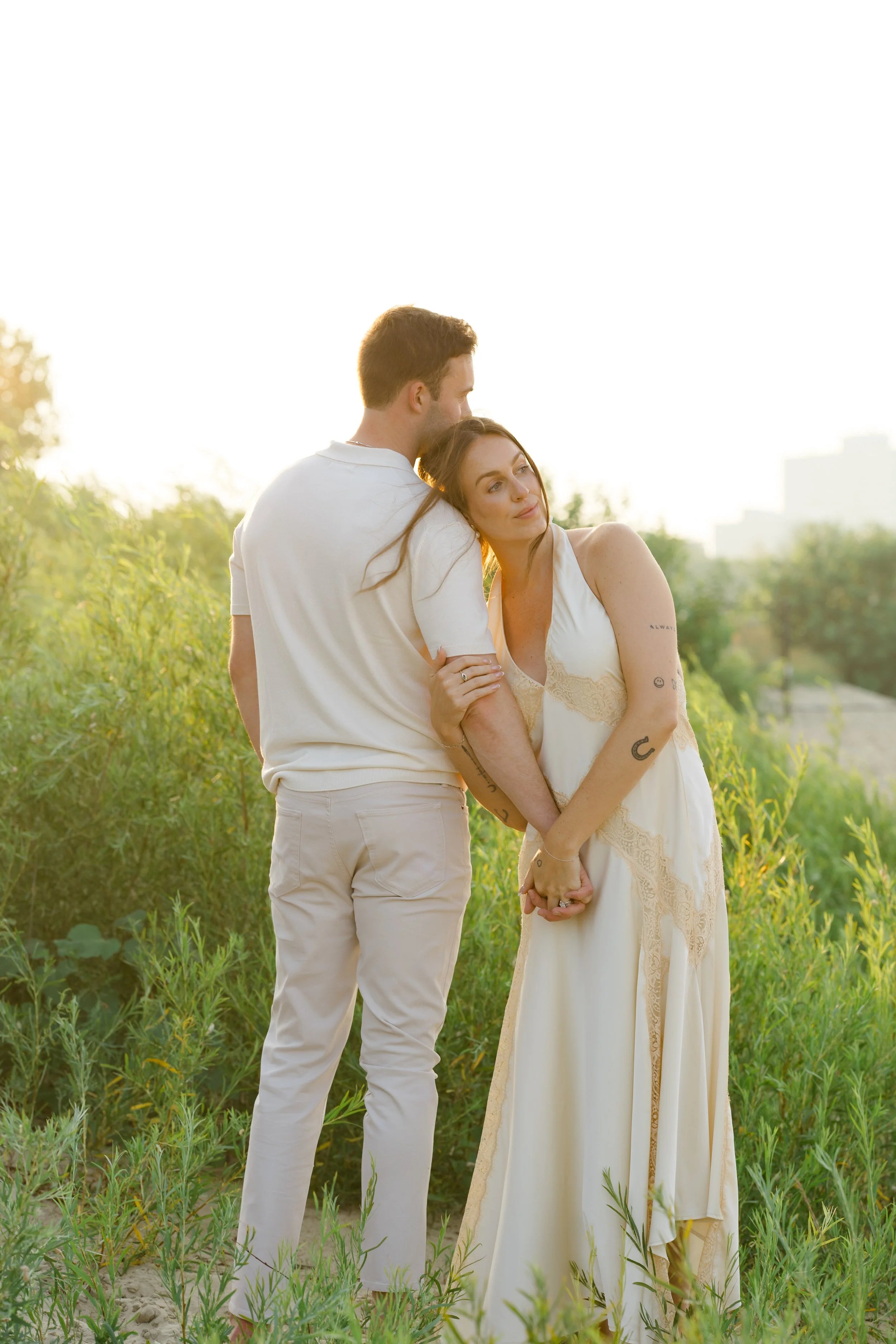 A man and woman holding hands in a grassy field with trees and a city skyline in the background, during sunset, embracing affectionately.