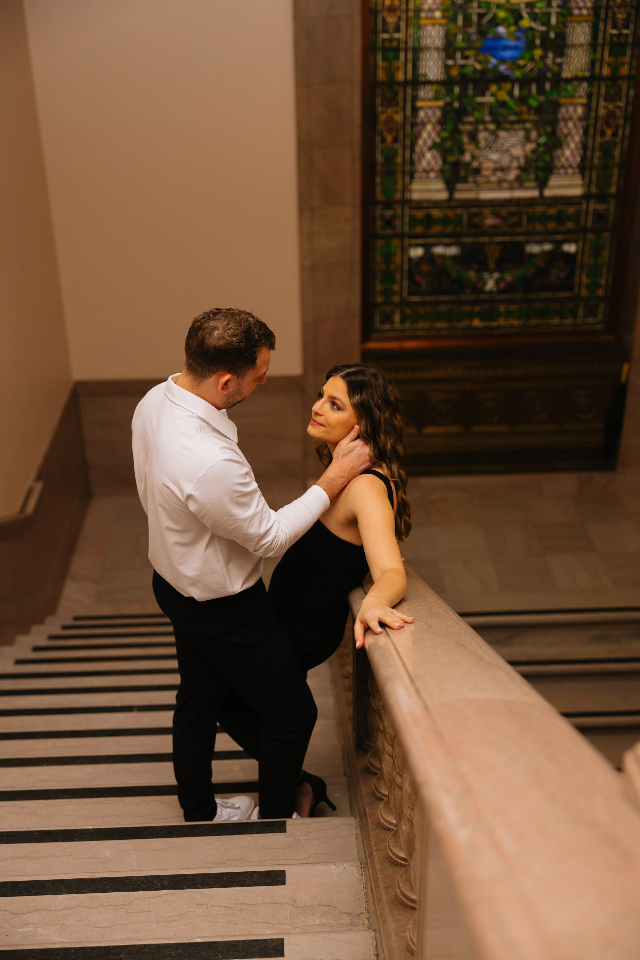 A man and a woman are on a staircase landing, the man holding the woman gently by the face and waist as they gaze into each other's eyes. The woman is sitting on the staircase railing with her hand resting on it, dressed in a black dress. The man is 