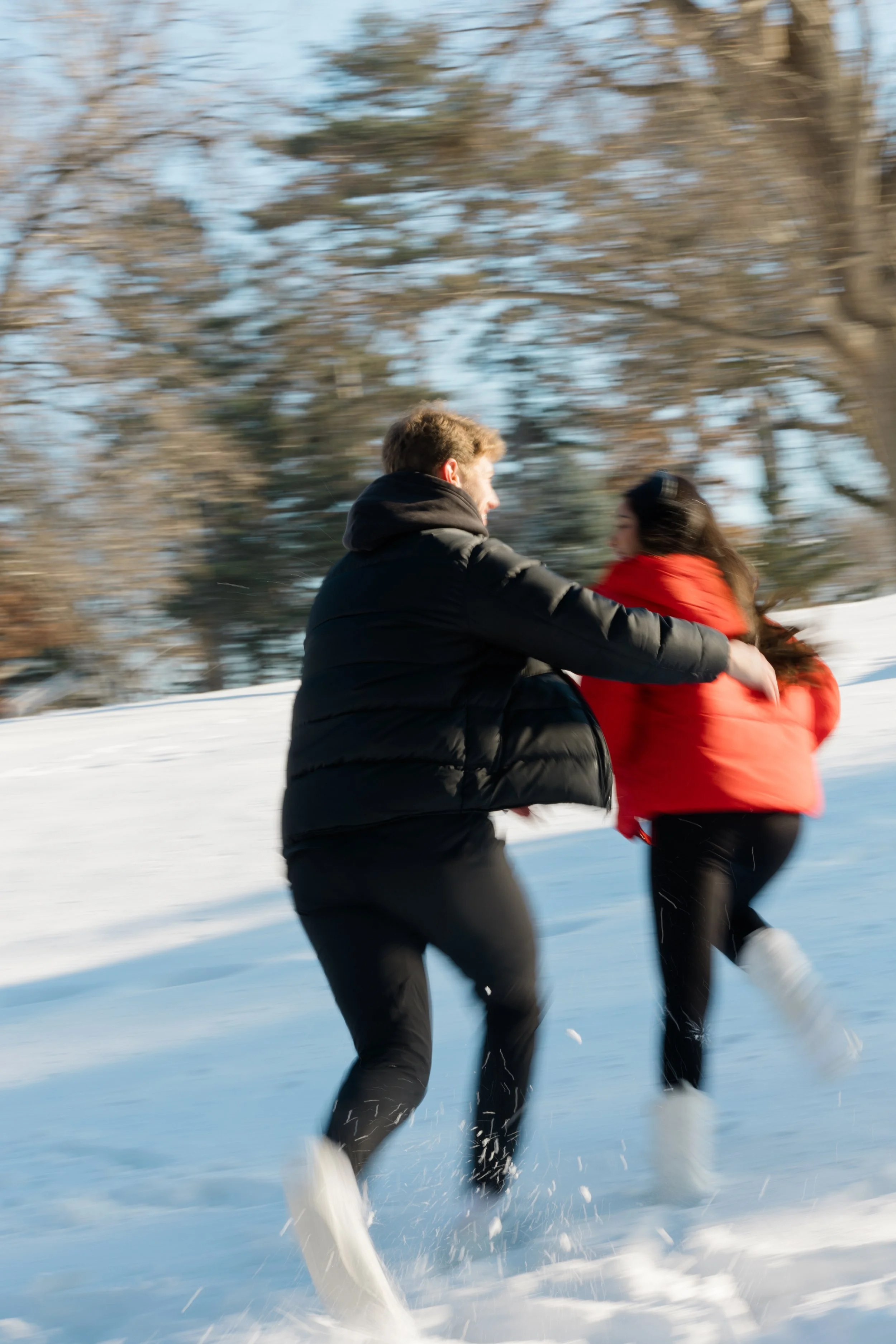 Two people in winter clothing running or playing in a snowy outdoor setting with blurred trees in the background.