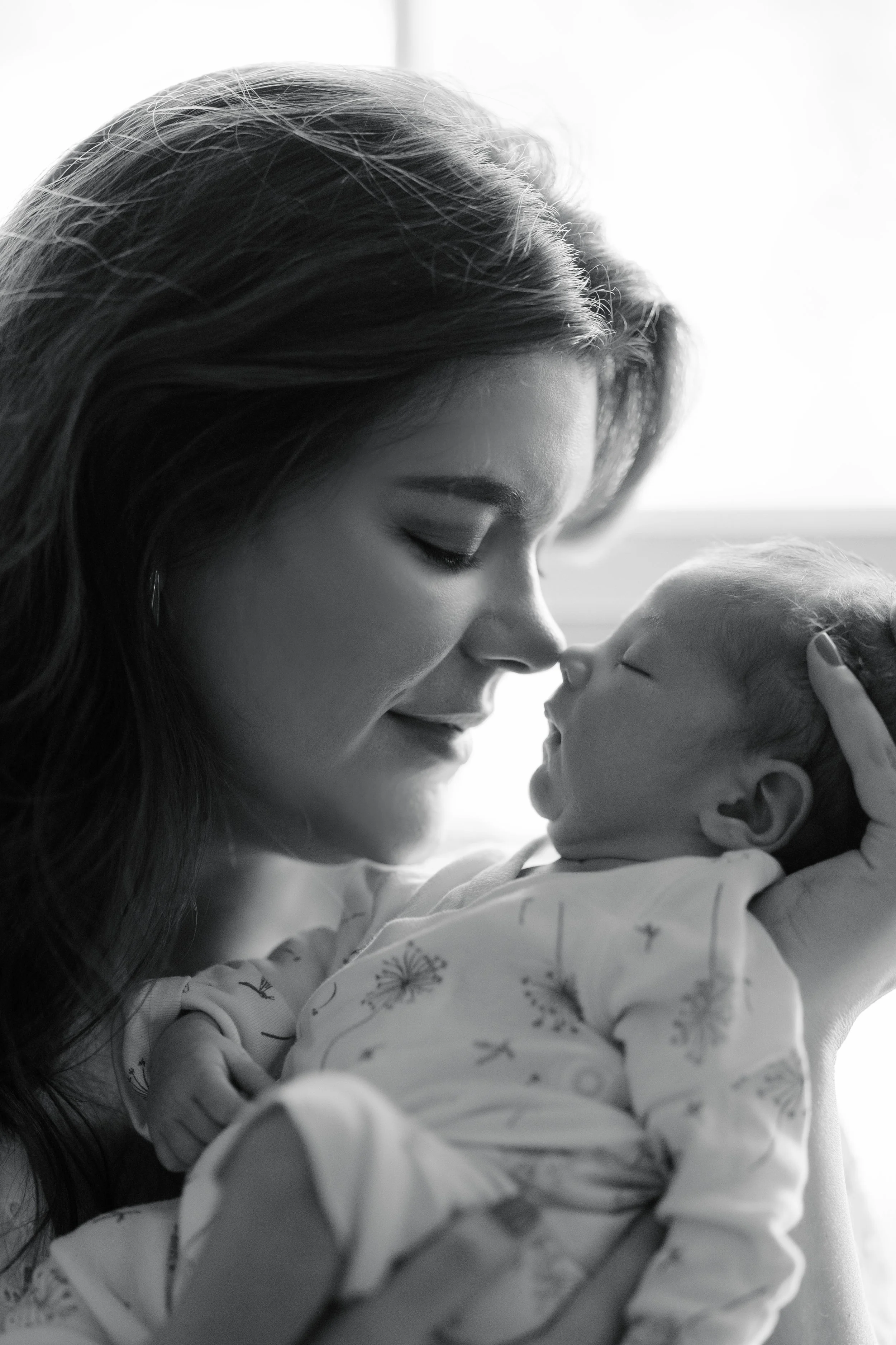 A woman with long hair and a patterned shirt gently touching noses with a sleeping baby, both in a close, intimate pose, with a soft background.
