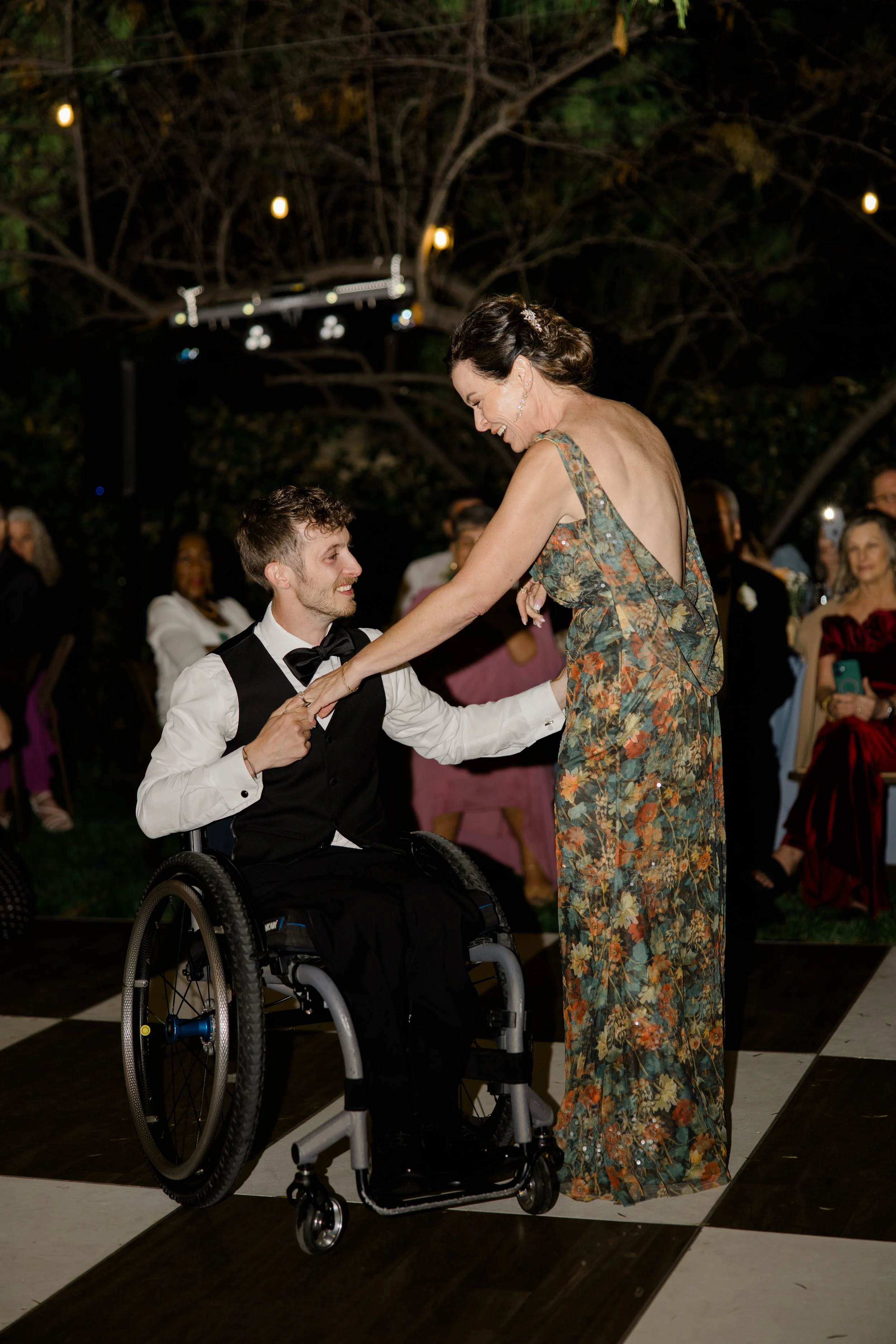 A man in a wheelchair and a woman in a floral dress dancing together at a celebration, with people in the background and string lights overhead.