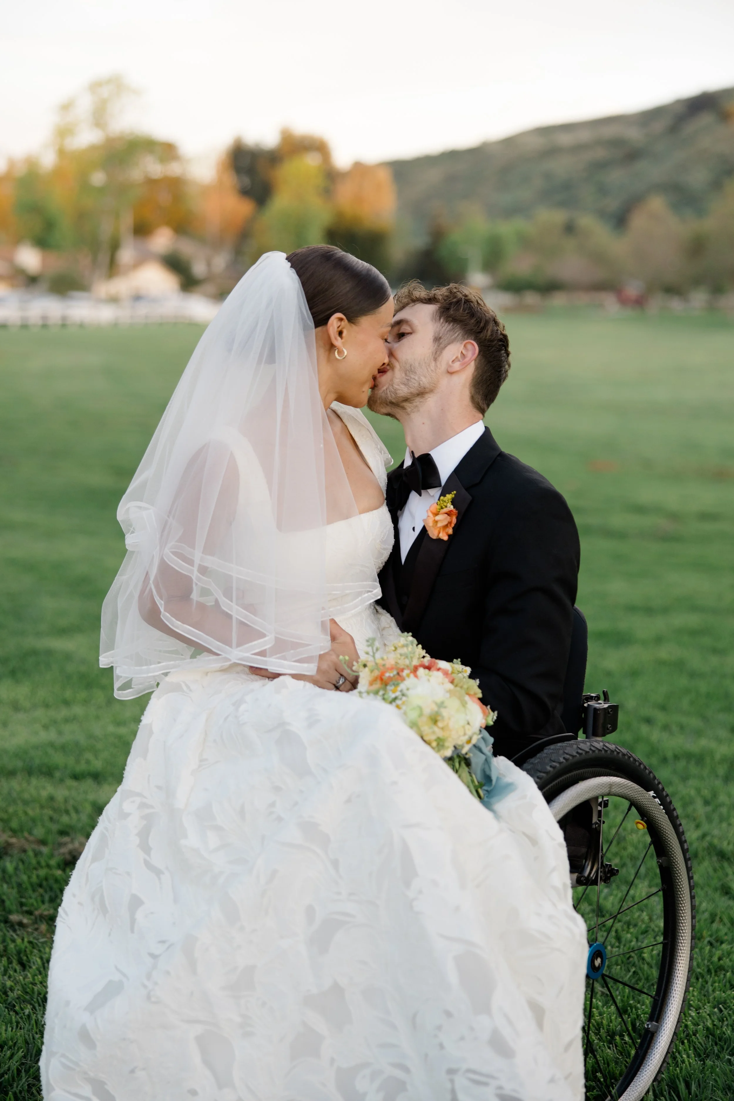 A bride and groom sharing a kiss outdoors on a grassy field with trees and hills in the background. The bride is wearing a white wedding dress and veil, holding a bouquet. The groom is in a black tuxedo with a boutonnière, and is seated in a wheelcha