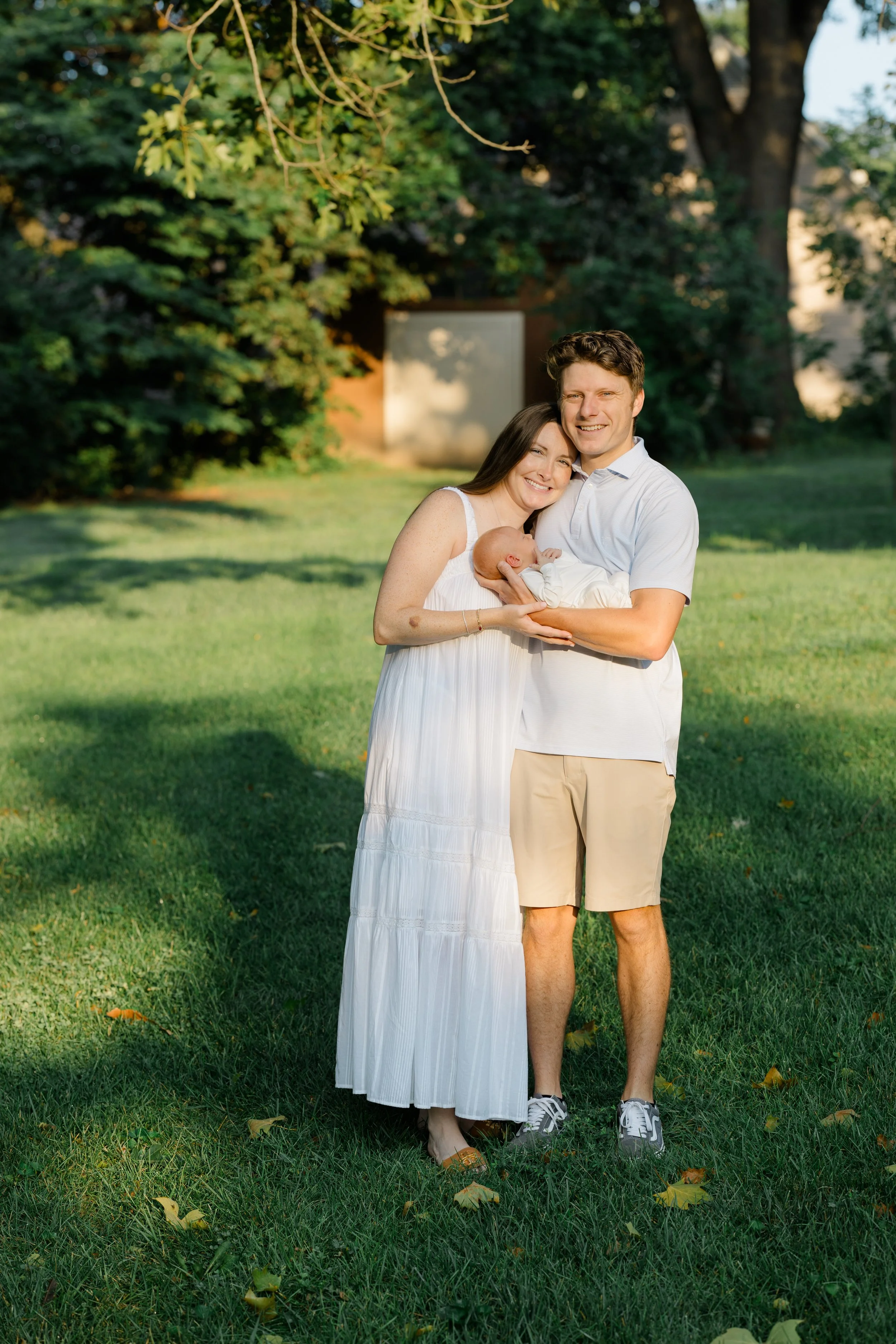 A happy family of three, a woman, a man, and a baby, standing outdoors in a park with green grass and trees, smiling and hugging, in warm, sunny weather.