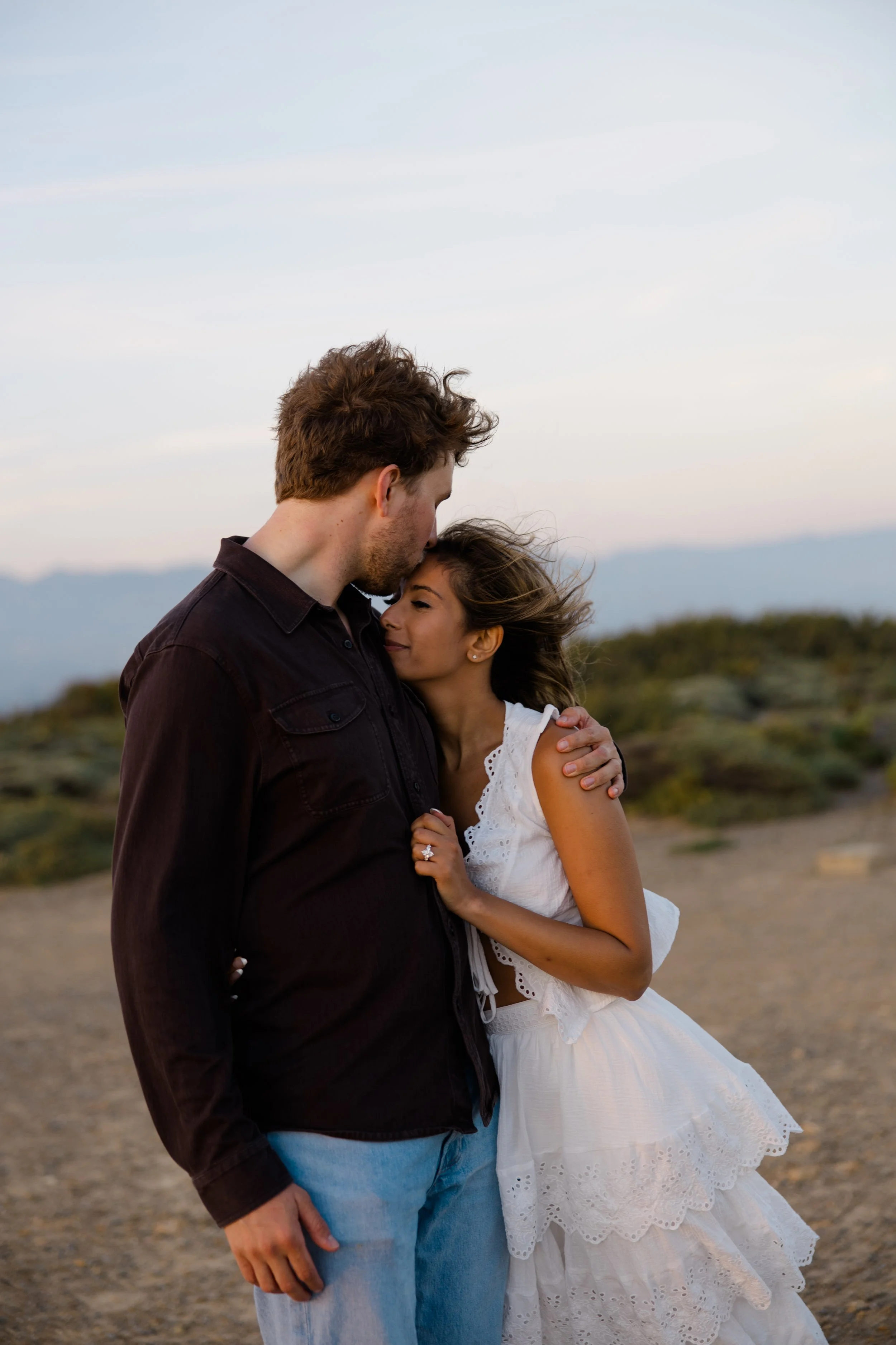 A couple embracing outdoors during sunset, with mountains in the background. The man, wearing a dark shirt and light blue jeans, is resting his forehead on the woman's forehead. The woman, wearing a white dress with lace details, is smiling with her 