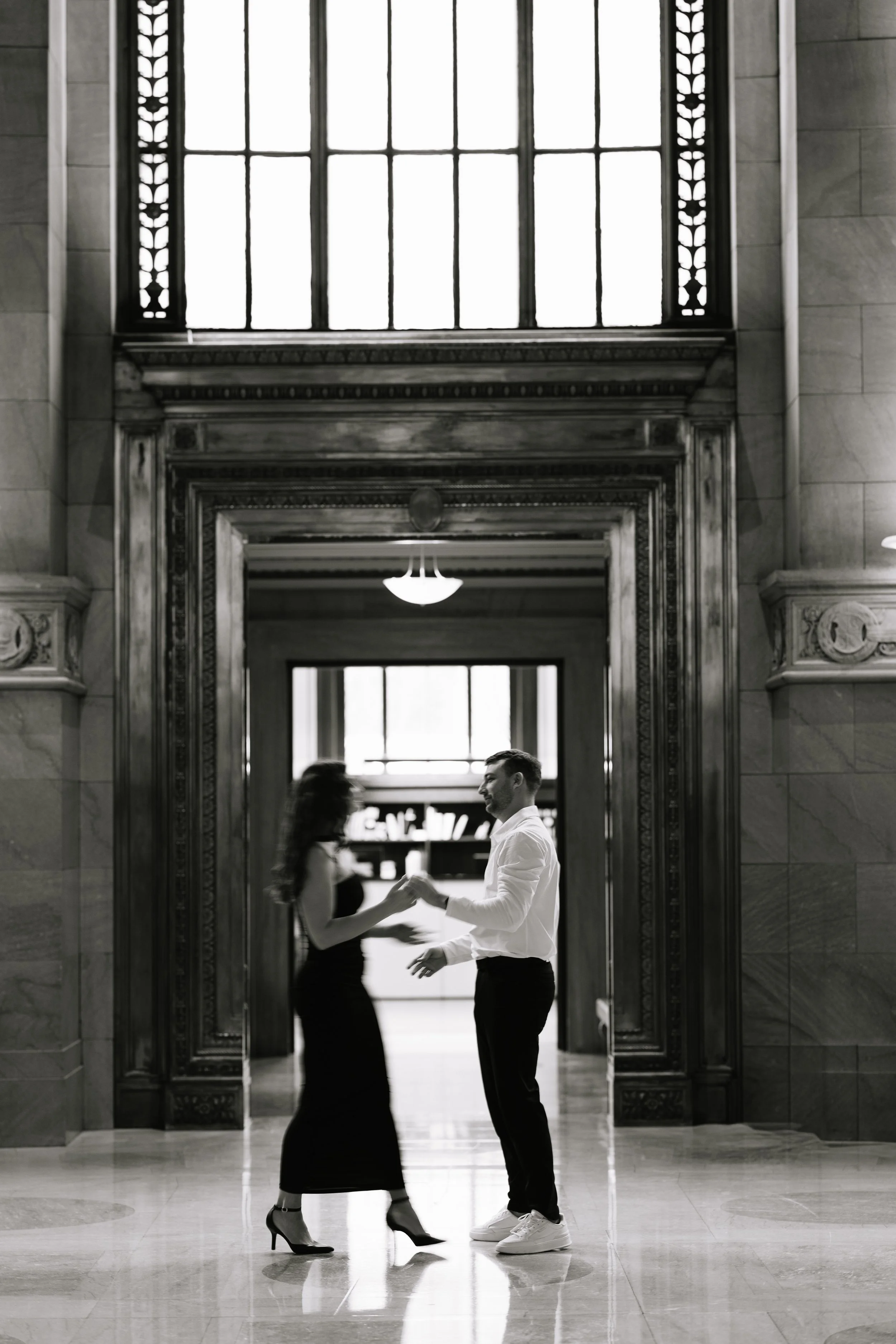 A man and woman dancing together in a grand hallway with ornate architectural details and large windows, black and white photograph.