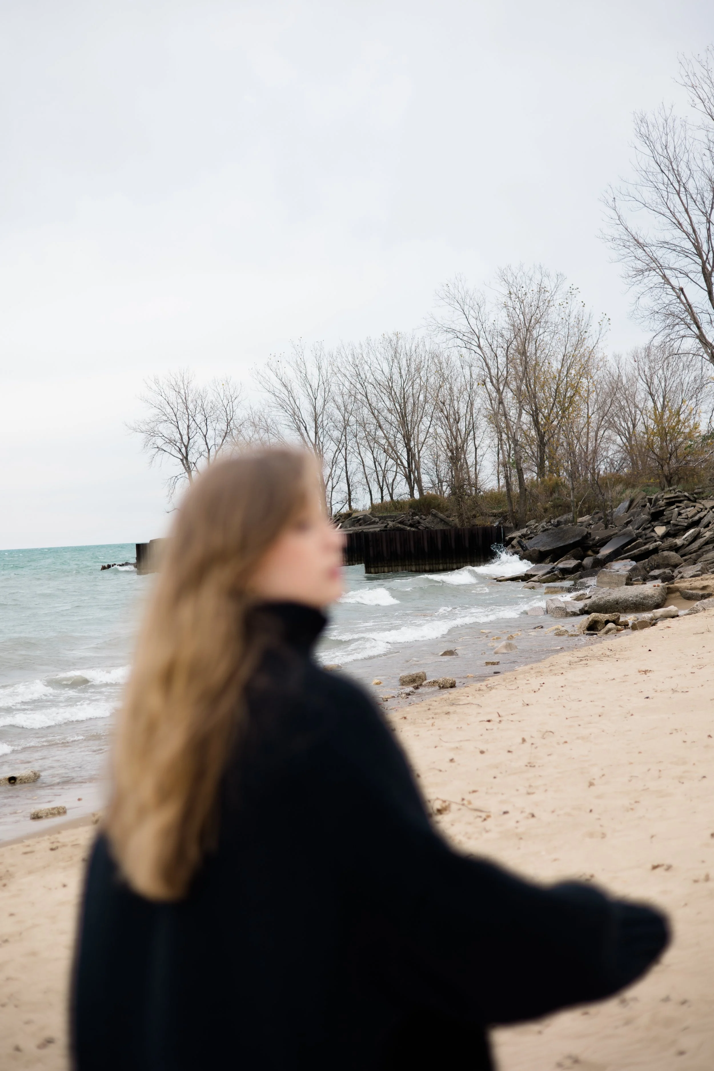 A woman with long blonde hair walking on a beach with cloudy sky, rocks, and leafless trees in the background.