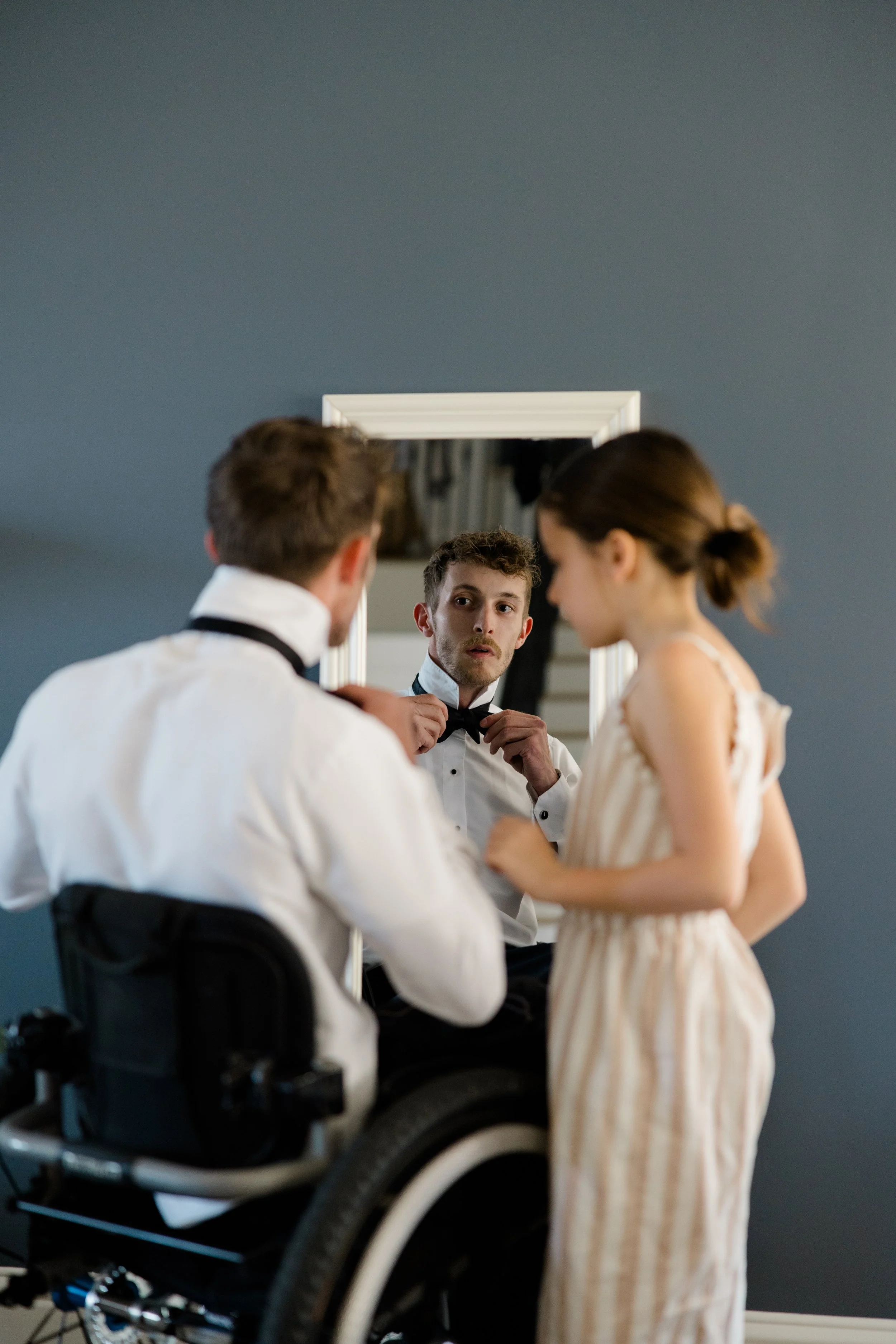 Young man in a tuxedo adjusting his bow tie while looking at a mirror, with a woman in a light dress standing beside him.