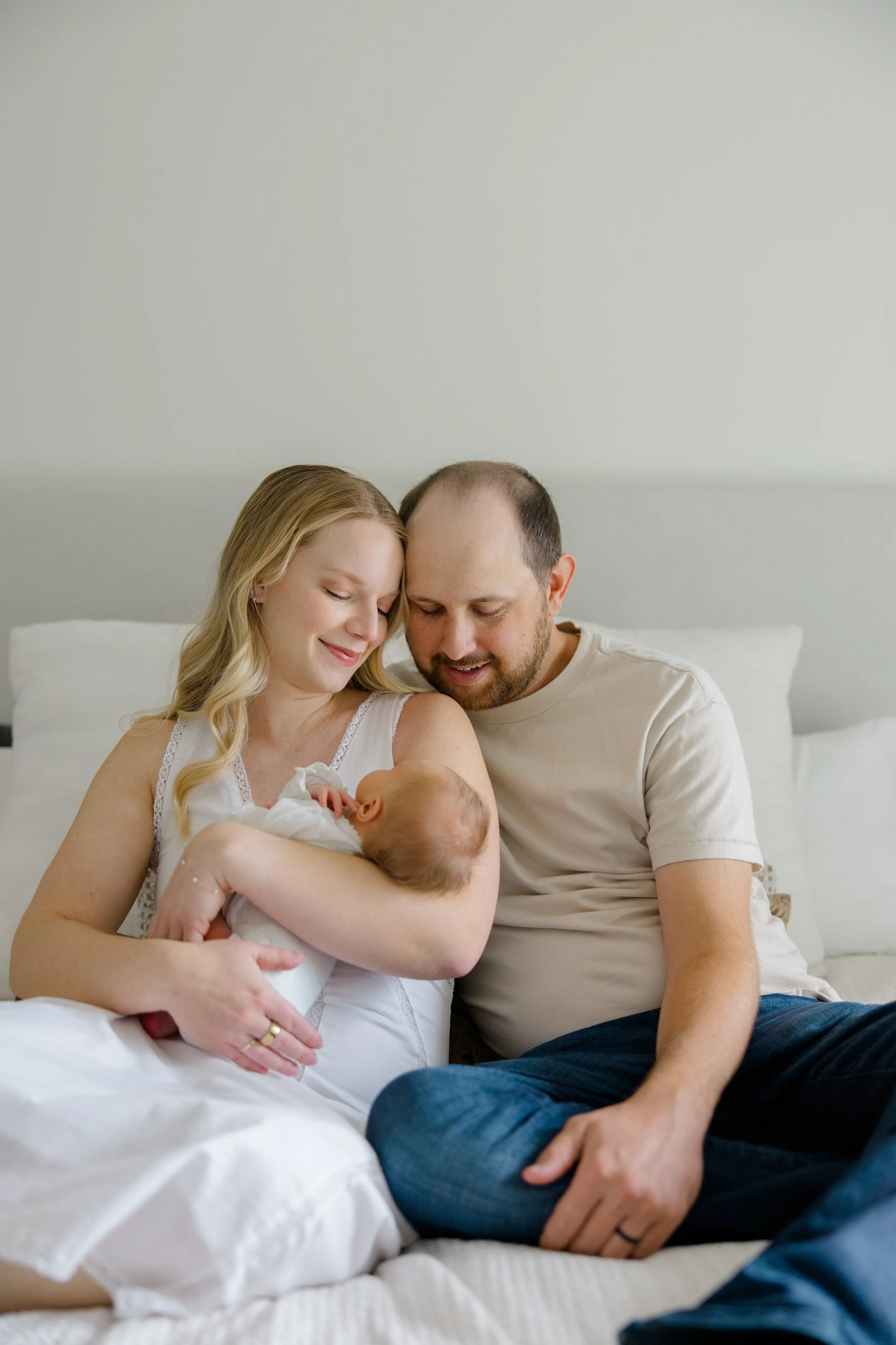 A mother and father holding and looking at a newborn baby in a bedroom with a bed and white walls.