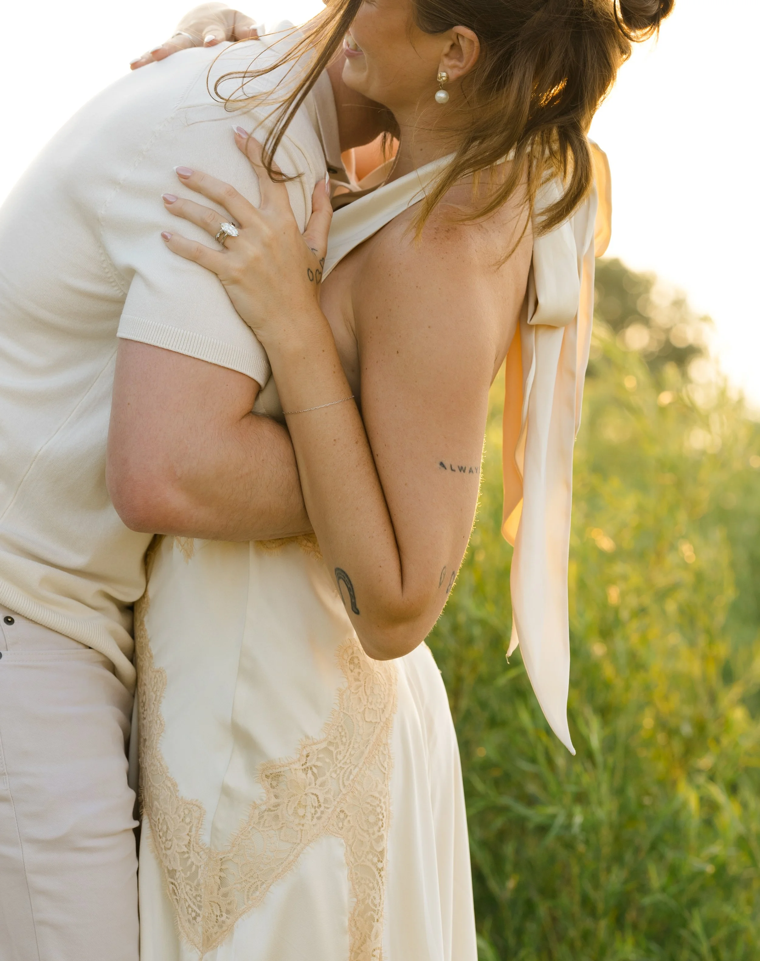 A person wearing a light-colored shirt and a wedding ring hugs and kisses another person dressed in a white dress with lace details, both outdoors during the golden hour.