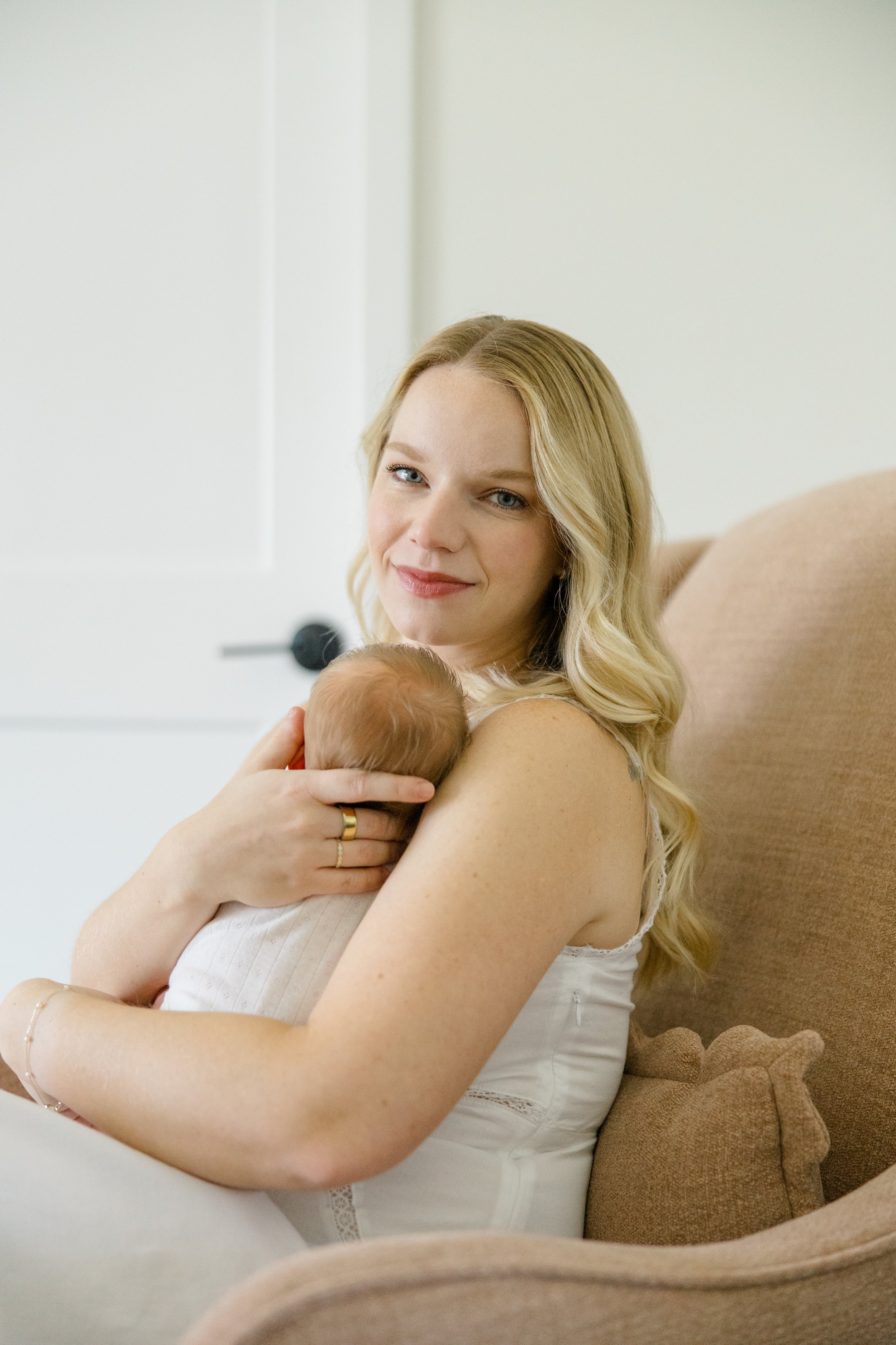 A young woman with long blonde hair and light skin holding a newborn baby, sitting on a beige couch in a bright room with white walls.