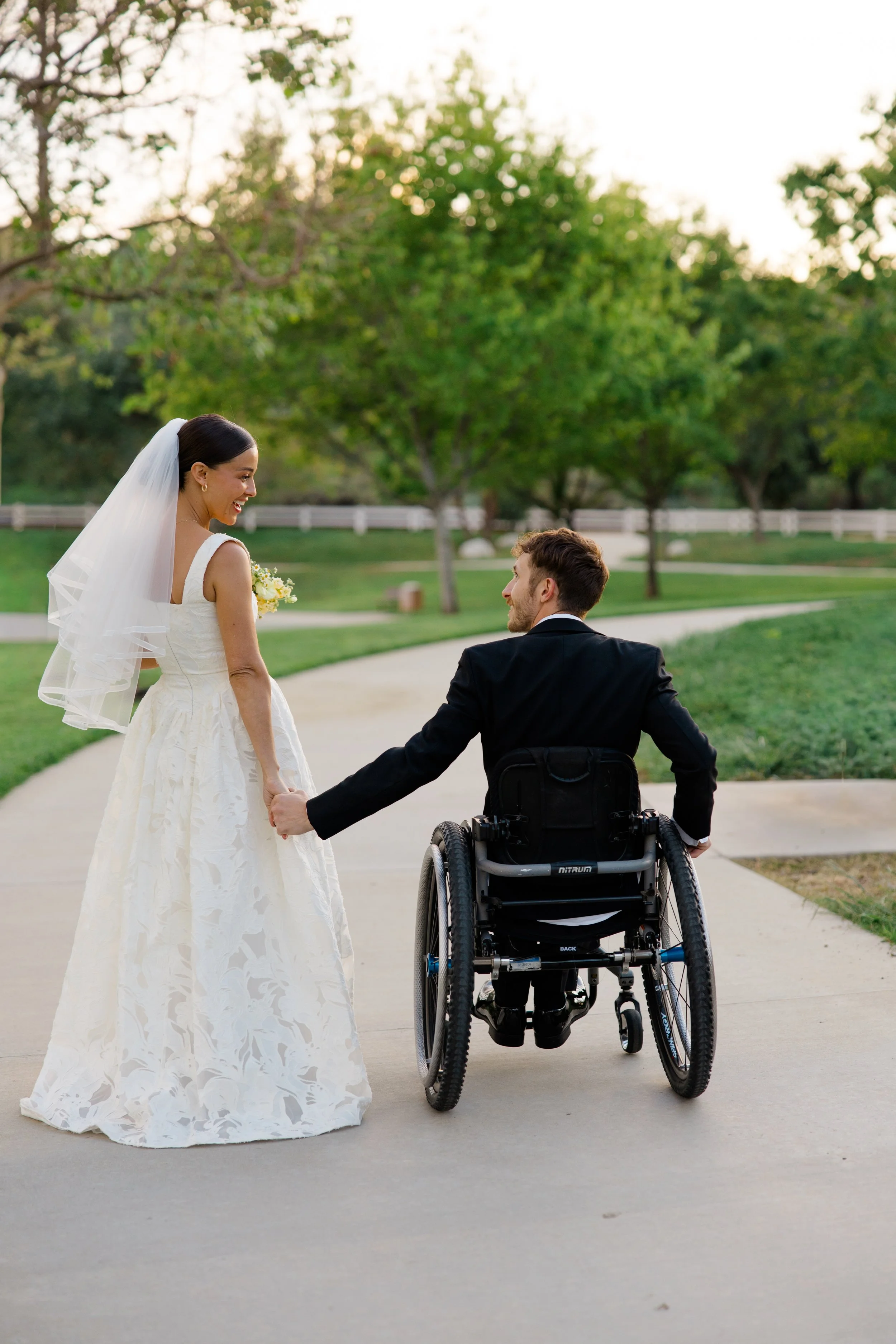 A bride in a white wedding dress and veil holds hands with a groom in a wheelchair on a paved path in a park with green trees and sunlight in the background.