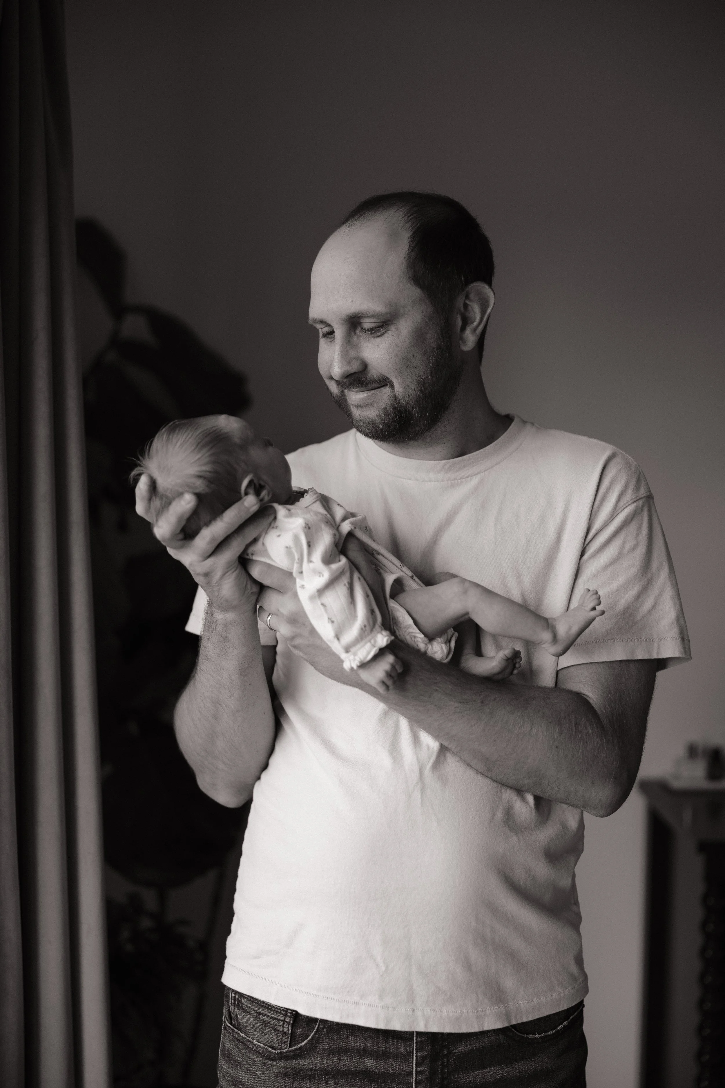 A man holding a newborn baby in a home setting, both facing each other and smiling. The scene is in black and white.