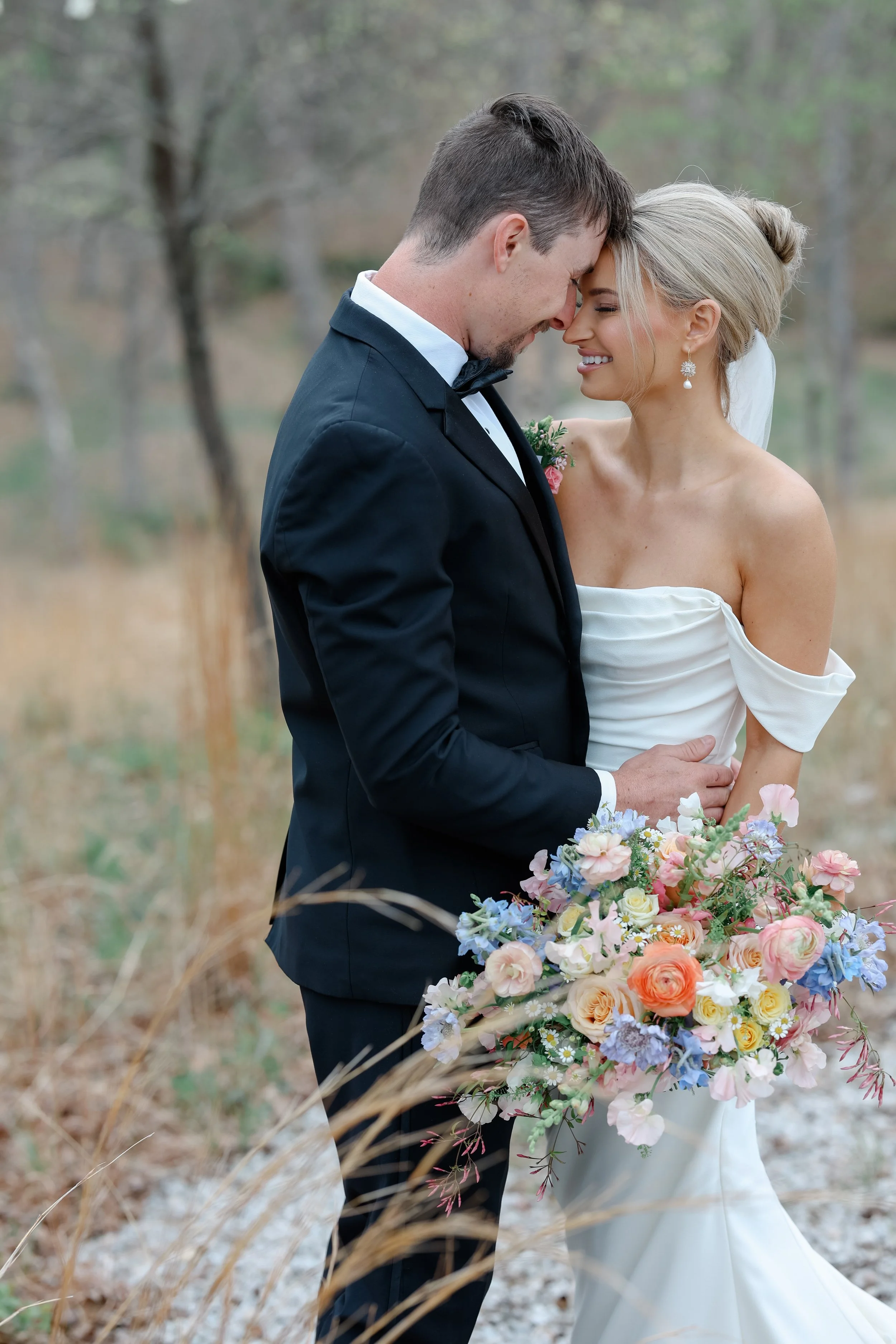 A bride and groom with their foreheads touching and eyes closed, standing outdoors with a blurred natural background, holding a large colorful bouquet of flowers.
