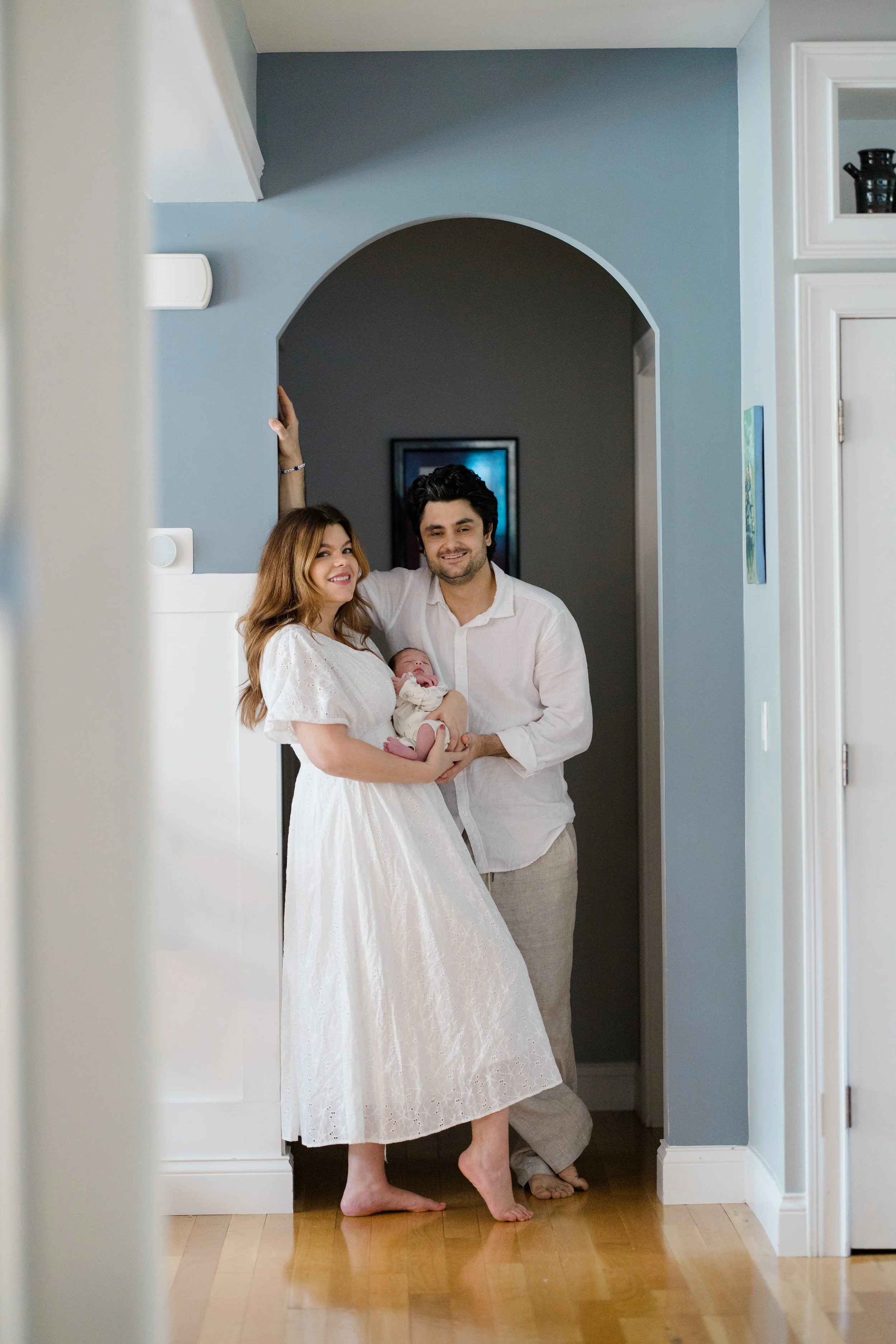 A family of three, including a woman in a white dress, a man, and a baby, standing in a doorway in a home with blue walls and hardwood floors.