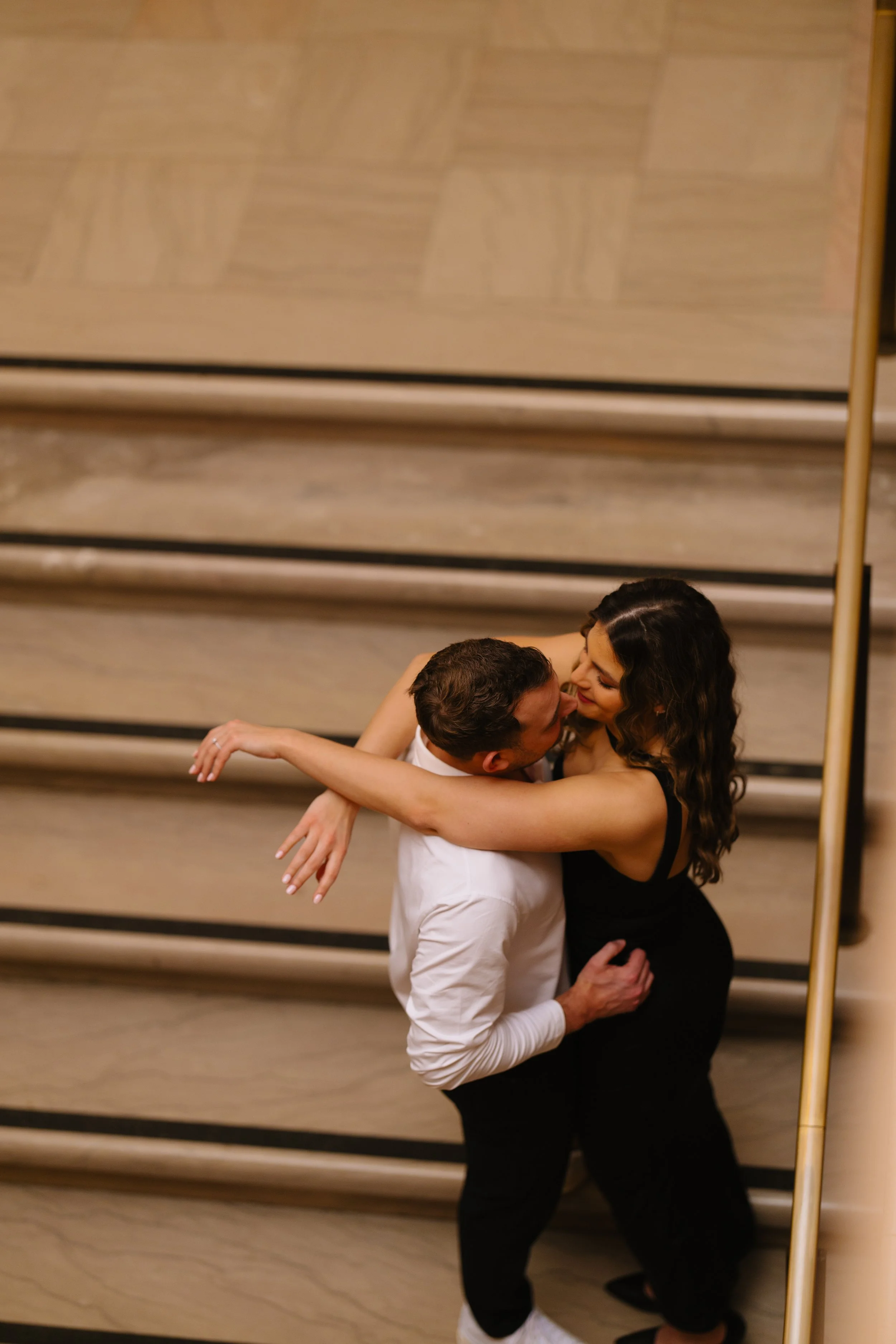 A couple dancing closely together in a dance studio with wooden walls and black horizontal lines. The woman has long dark hair and is wearing a black dress, and the man has short dark hair and is dressed in a white shirt and black pants.