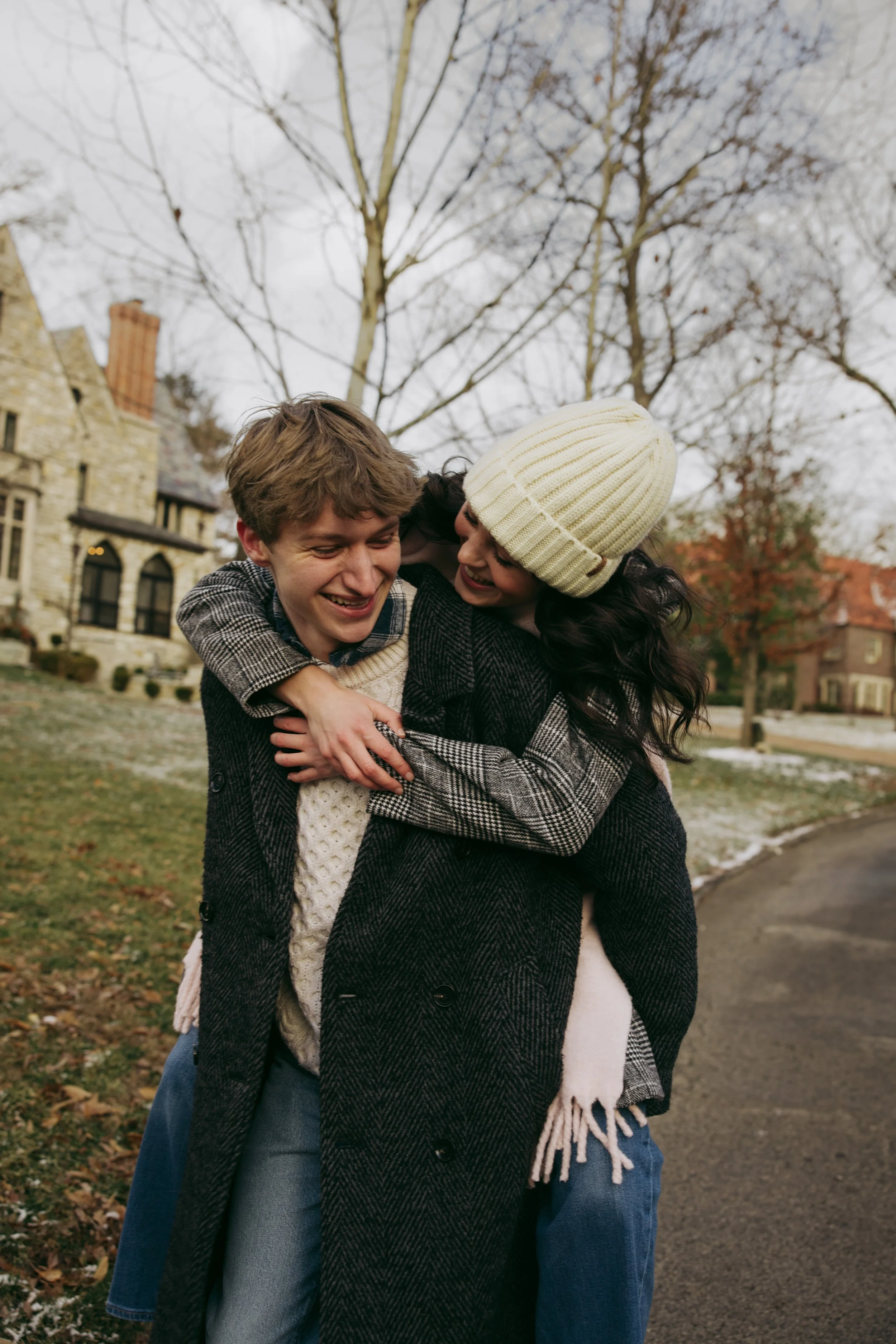 Two young women hugging outdoors in a park with leafless trees and houses in the background during autumn.