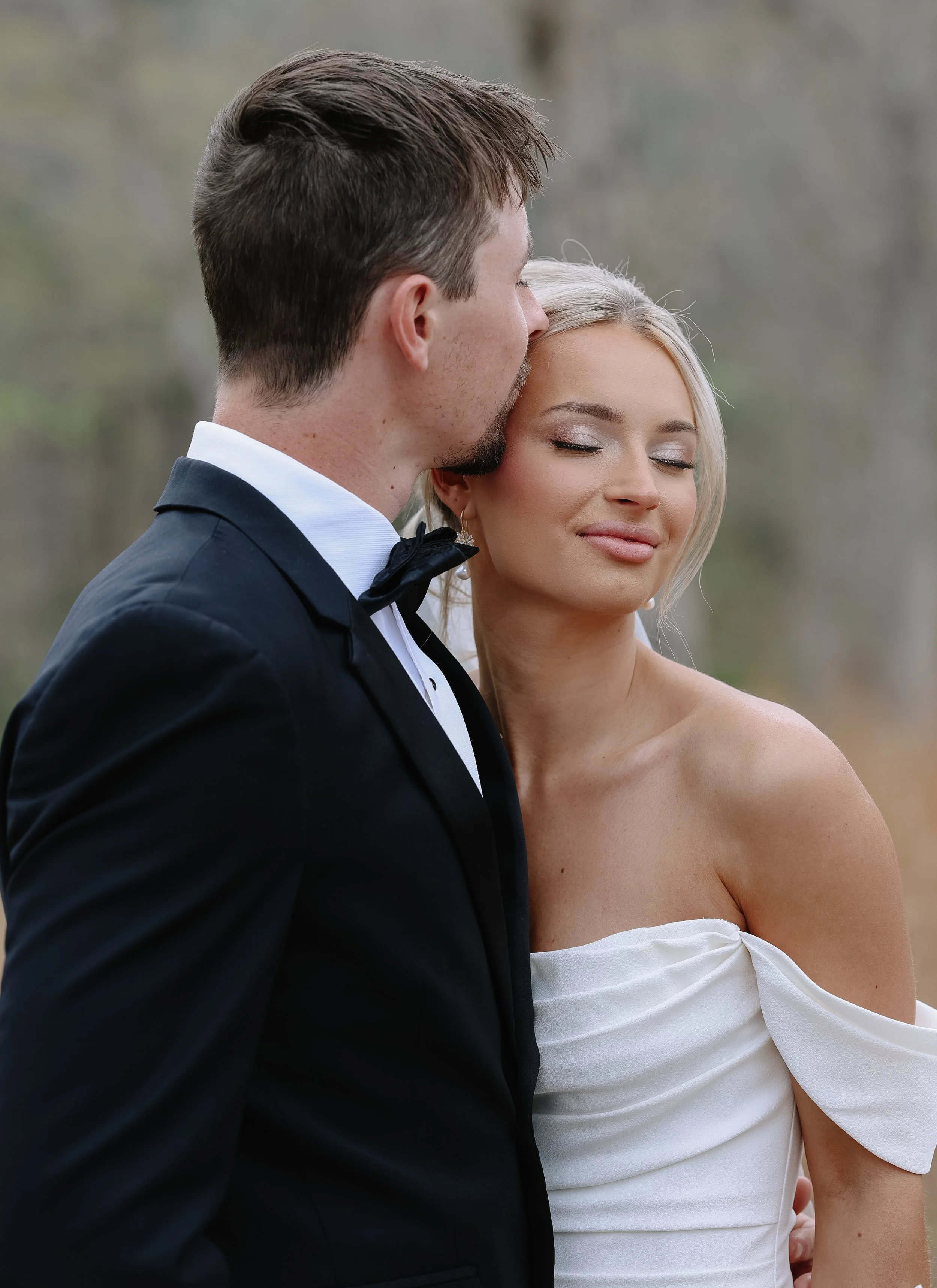 A man in a tuxedo with a bow tie kissing a woman in a strapless white dress with a silver earring.